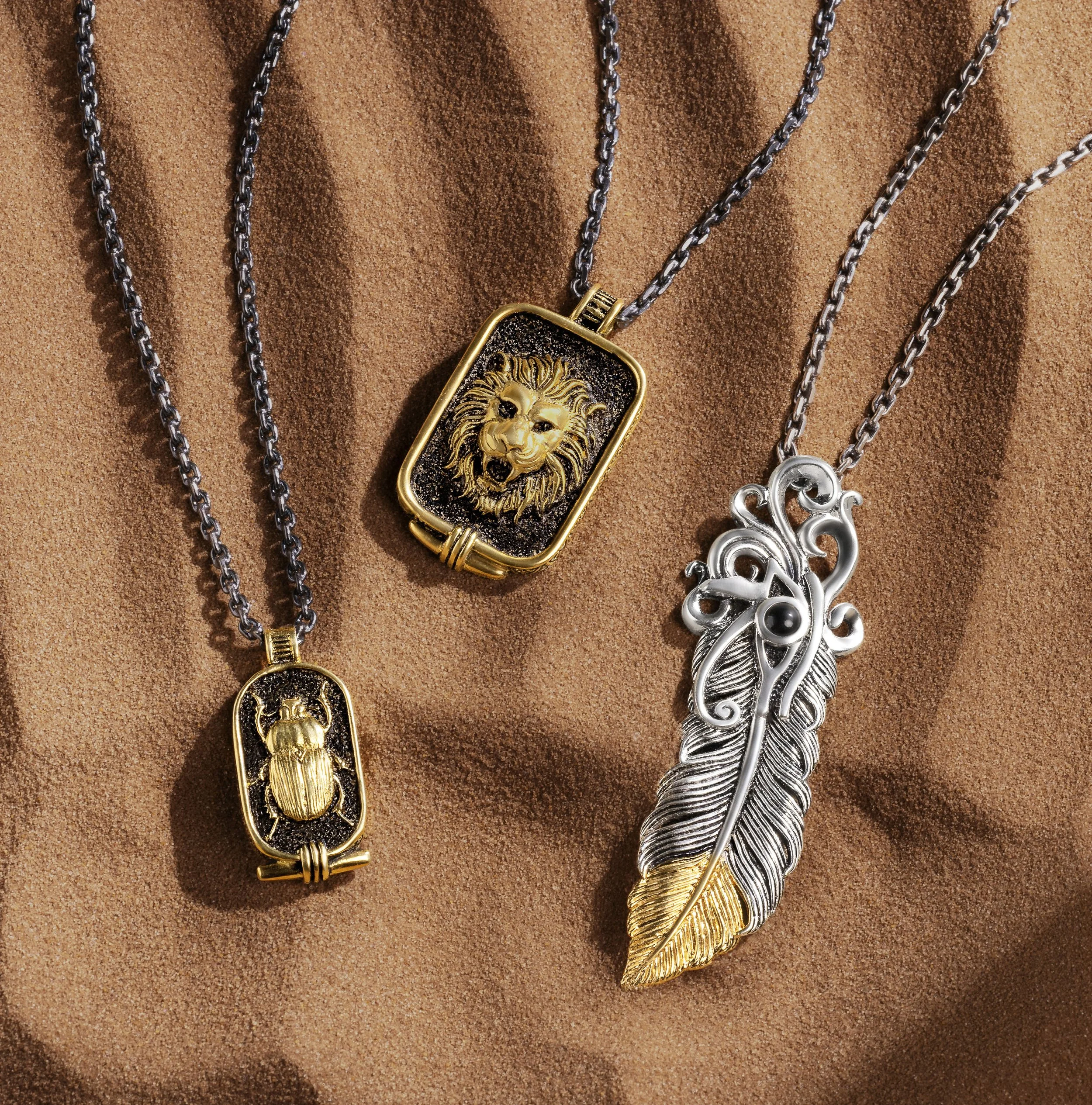 Three necklaces with animal-themed pendants: a lion, a beetle, and a feather, displayed on a brown textured fabric background.
