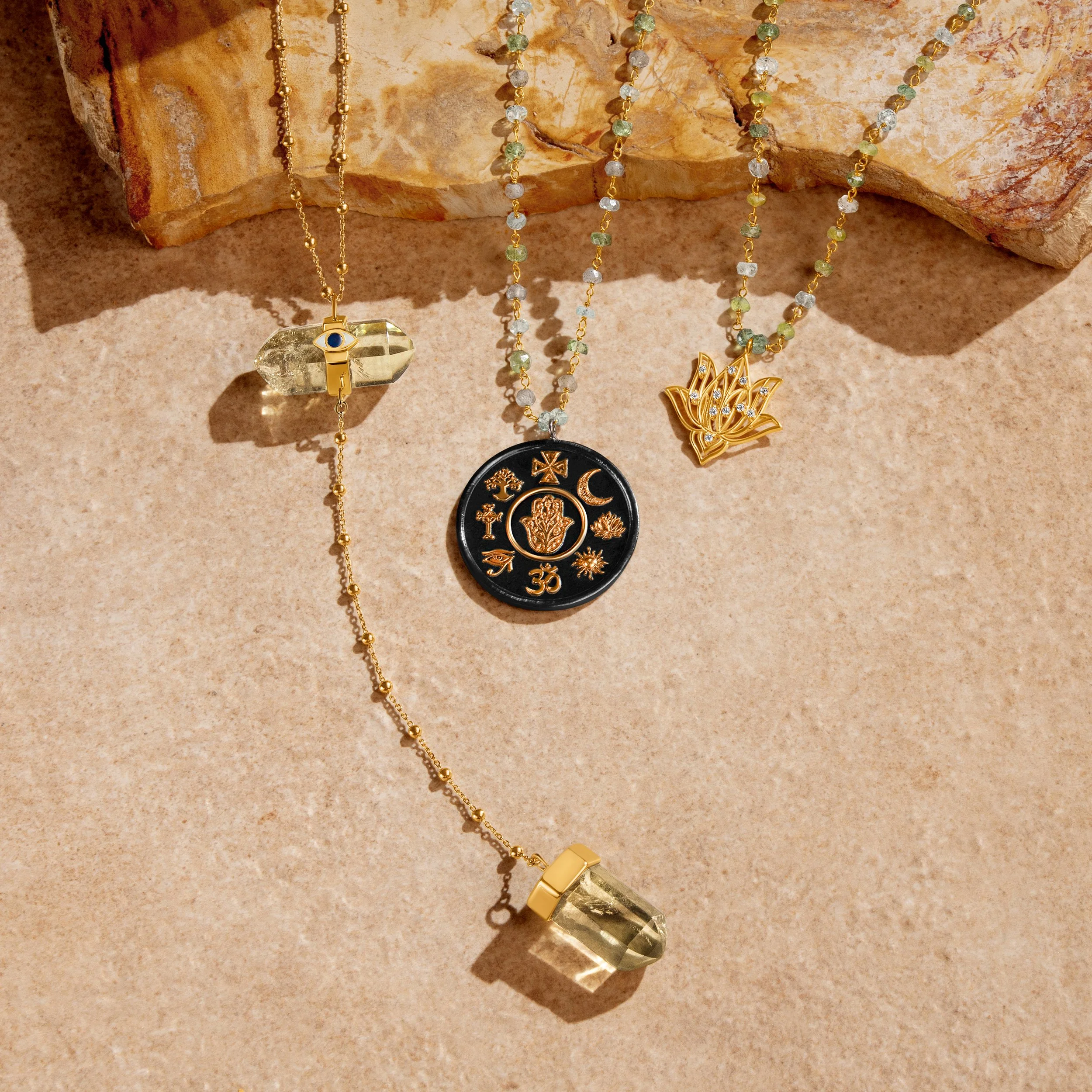Three gold necklaces with pendants, including a lotus flower, religious symbols, and a gemstone, displayed on a beige surface with a rock in the background.