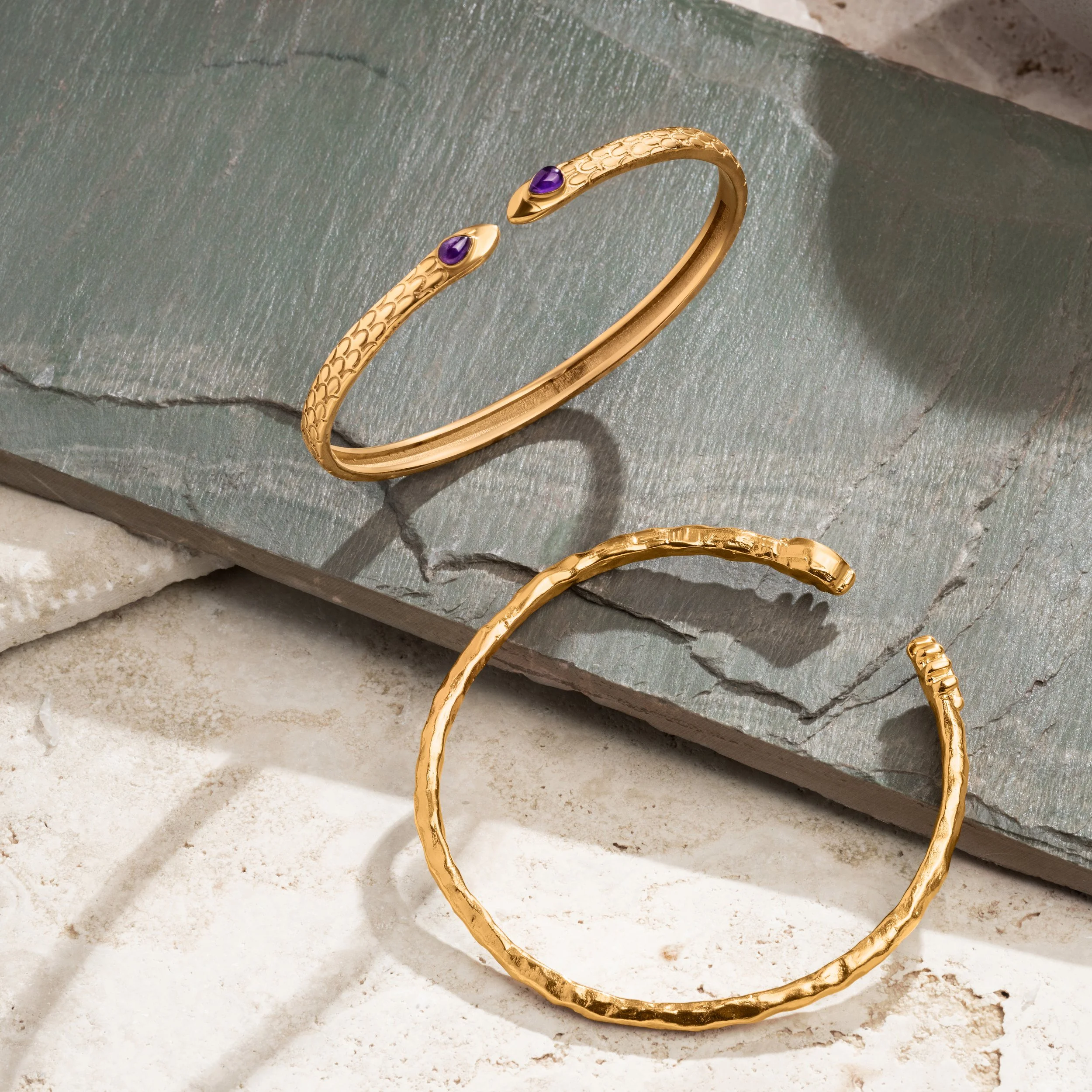 Gold bracelet with purple gemstones and textured gold bangles on a stone surface.