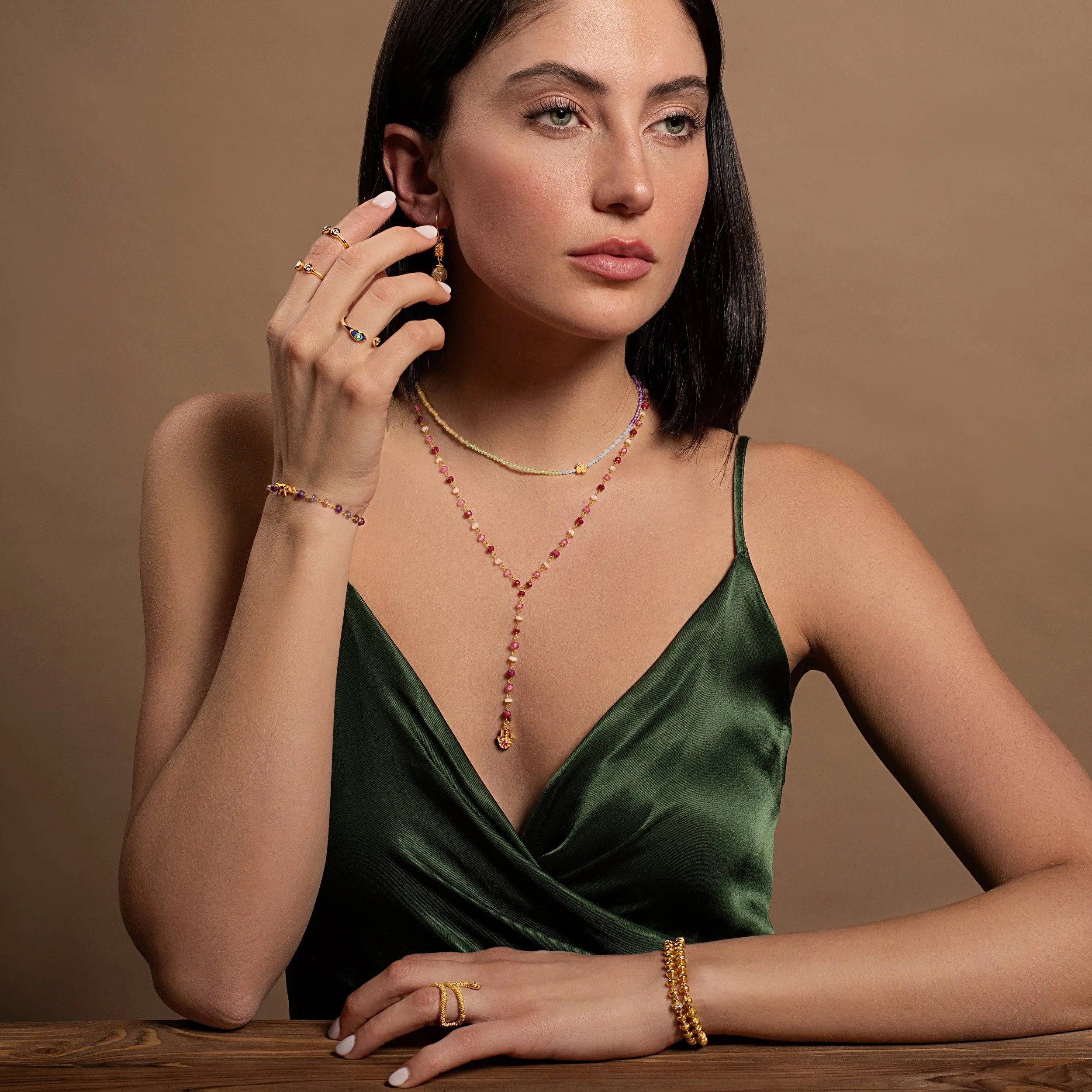 A woman with dark hair wearing a green satin dress and gold jewelry, including rings, necklaces, earrings, bracelets, and a ring on her finger, sitting at a wooden table against a plain background.