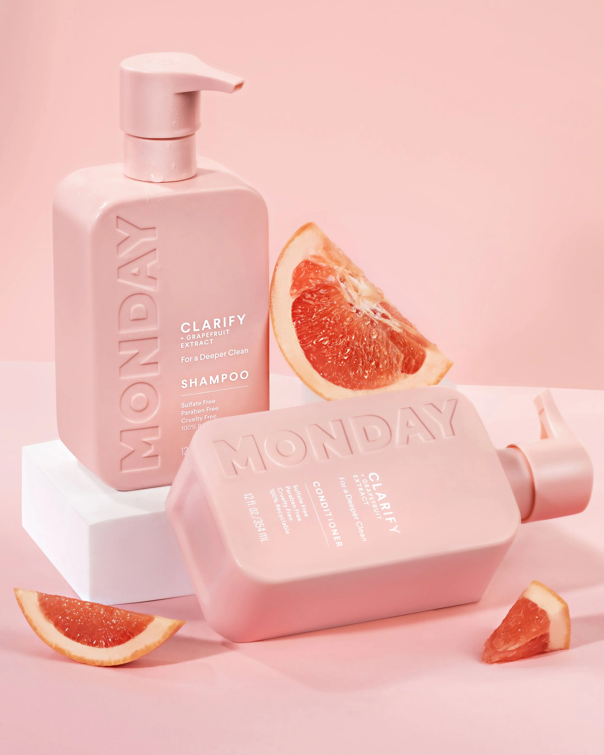 Pink bottles of shampoo and conditioner with embossed text 'MONDAY' and 'MONDAY,' accompanied by slices of grapefruit on a pink background.