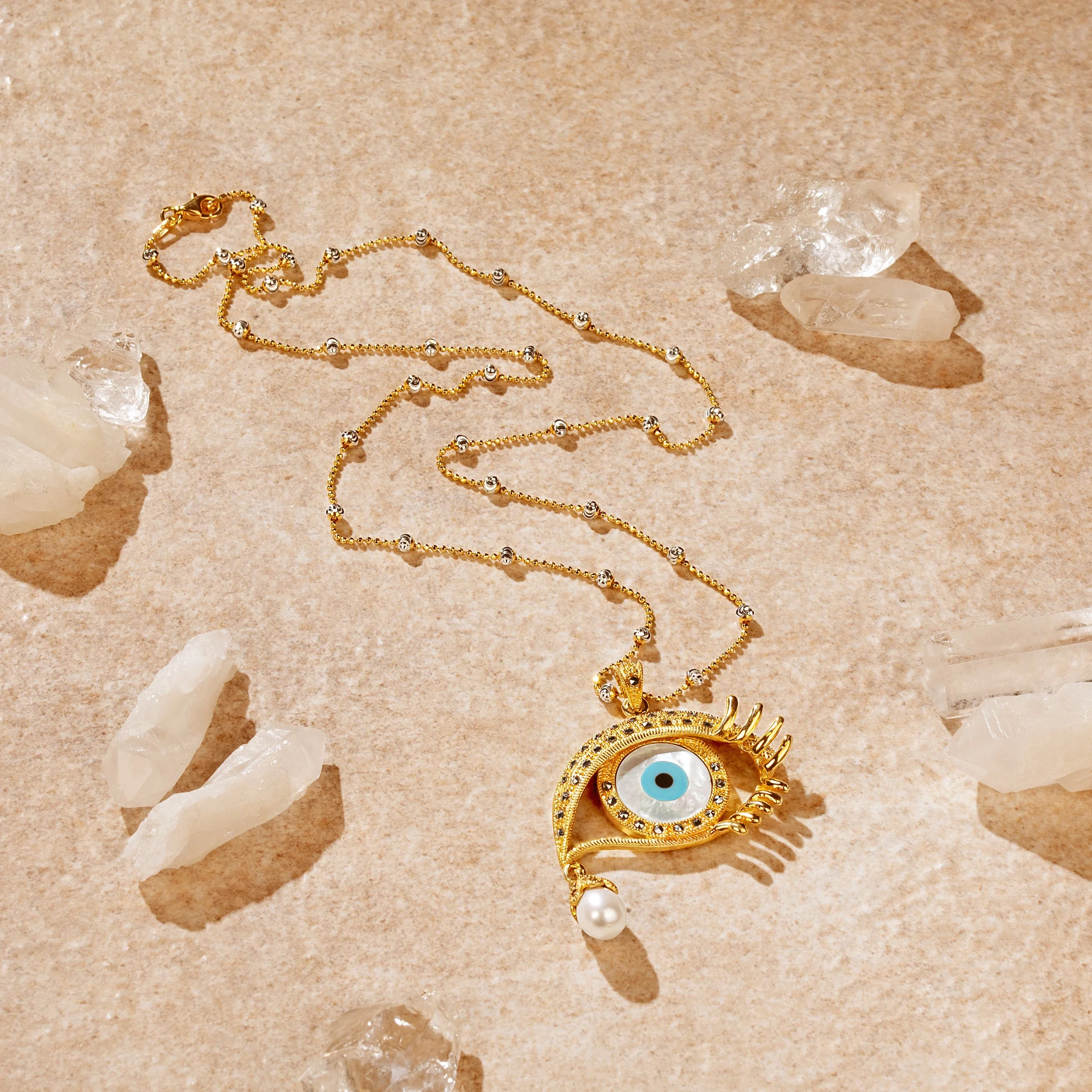 Gold necklace with eye-shaped pendant featuring a blue eye and pearl detail, surrounded by clear crystals, on a beige surface.