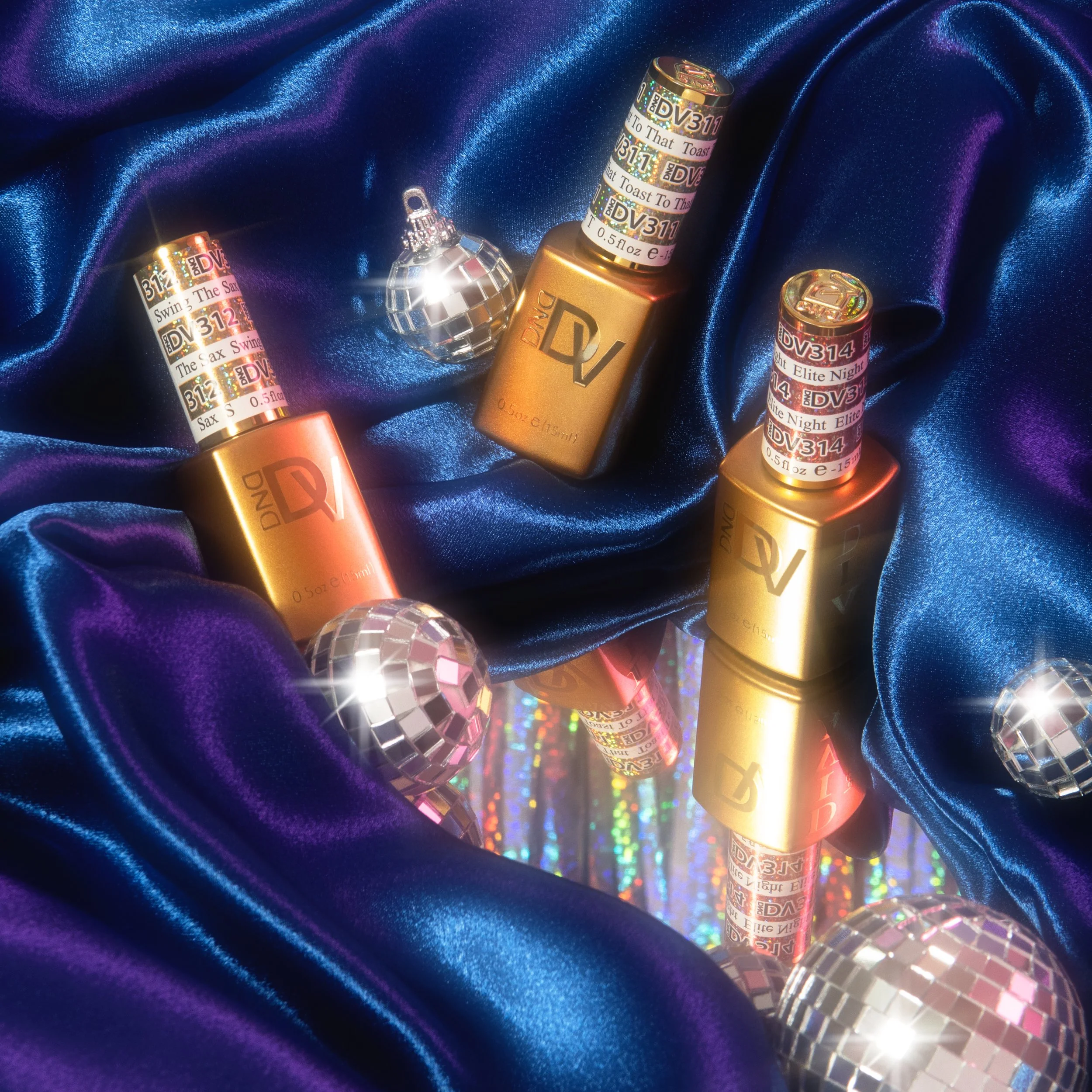 Three metallic lipstick tubes with holographic labels on a shiny blue satin fabric, along with mirrored disco balls reflecting light and colorful rainbow patterns.