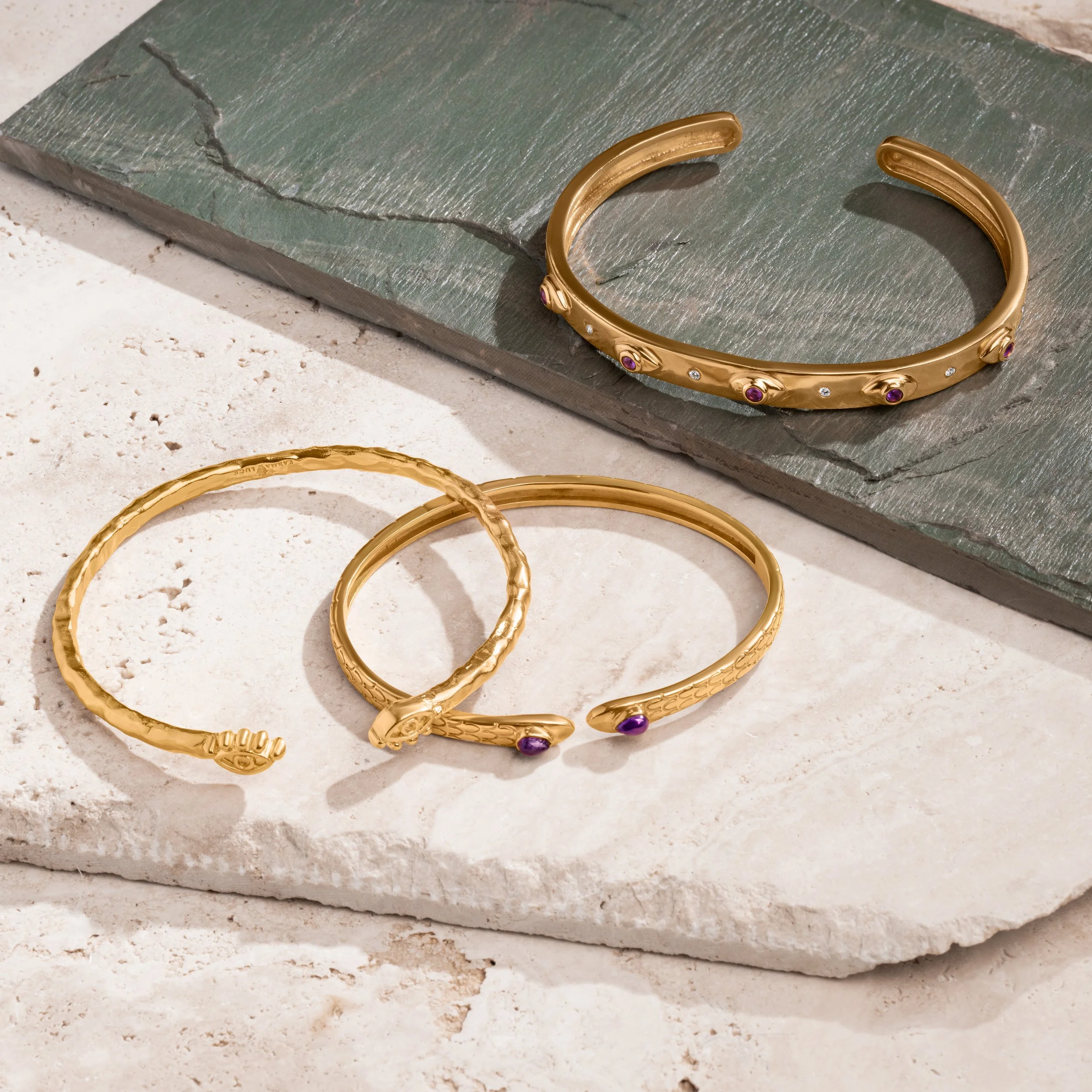 Three gold jewelry bracelets with purple and clear gemstones on a stone surface, with a gray slate tile in the background.