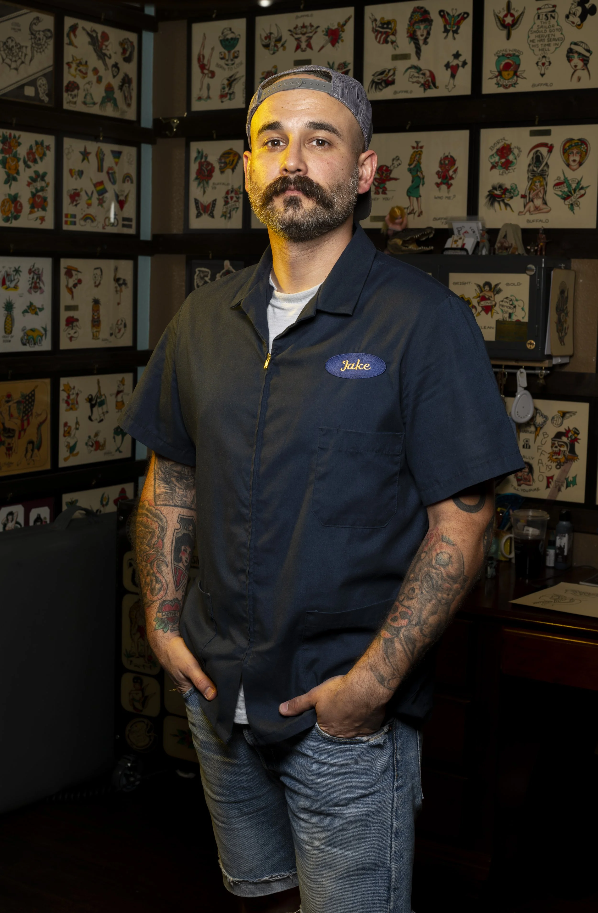 A man with a beard, tattoos, and a backwards baseball cap stands in front of tattoo flash art on the wall. He is wearing a navy blue work shirt with a name tag that says 'Jake' and light blue jeans.