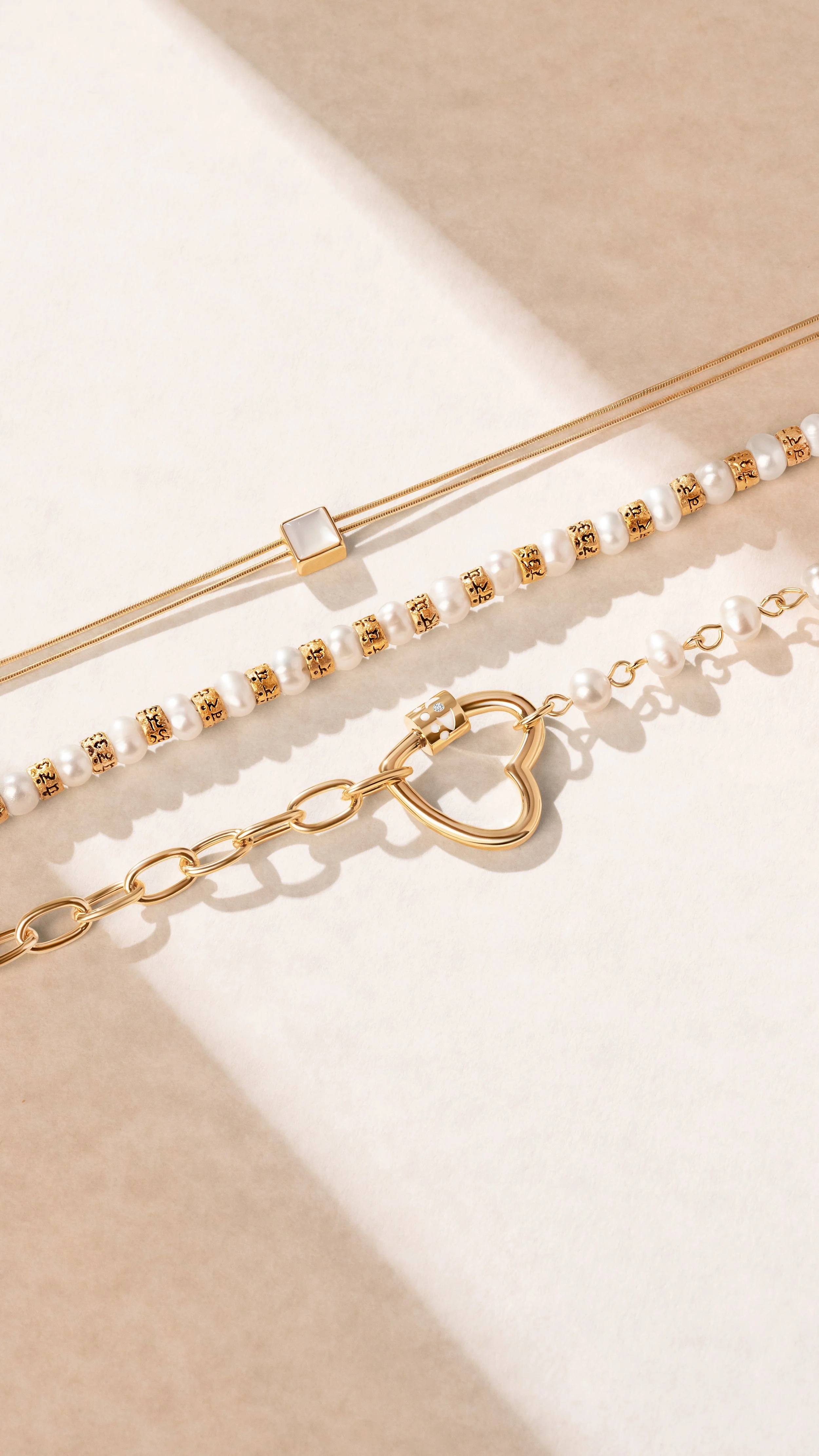 Three pieces of jewelry - a thin gold necklace with a square white pendant, a pearl and gold beaded bracelet, and a gold chain bracelet with a heart-shaped charm and small pearl accents - laid out on a beige surface with shadows.