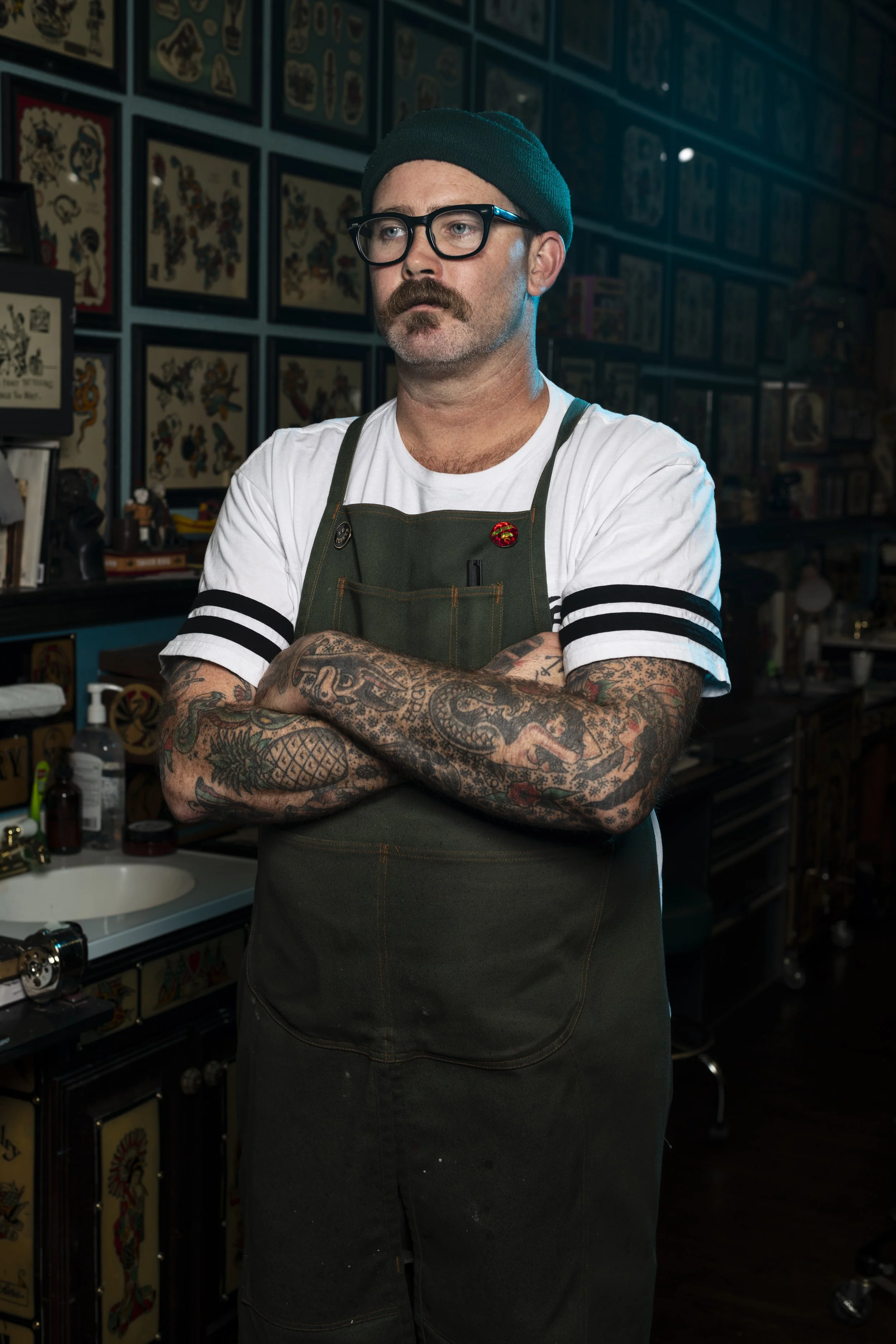 A man with tattoos on his arms, wearing glasses, a black beanie, a white t-shirt with black striped sleeves, and a green apron, stands with arms crossed inside a tattoo shop decorated with framed tattoo flash art on the walls.