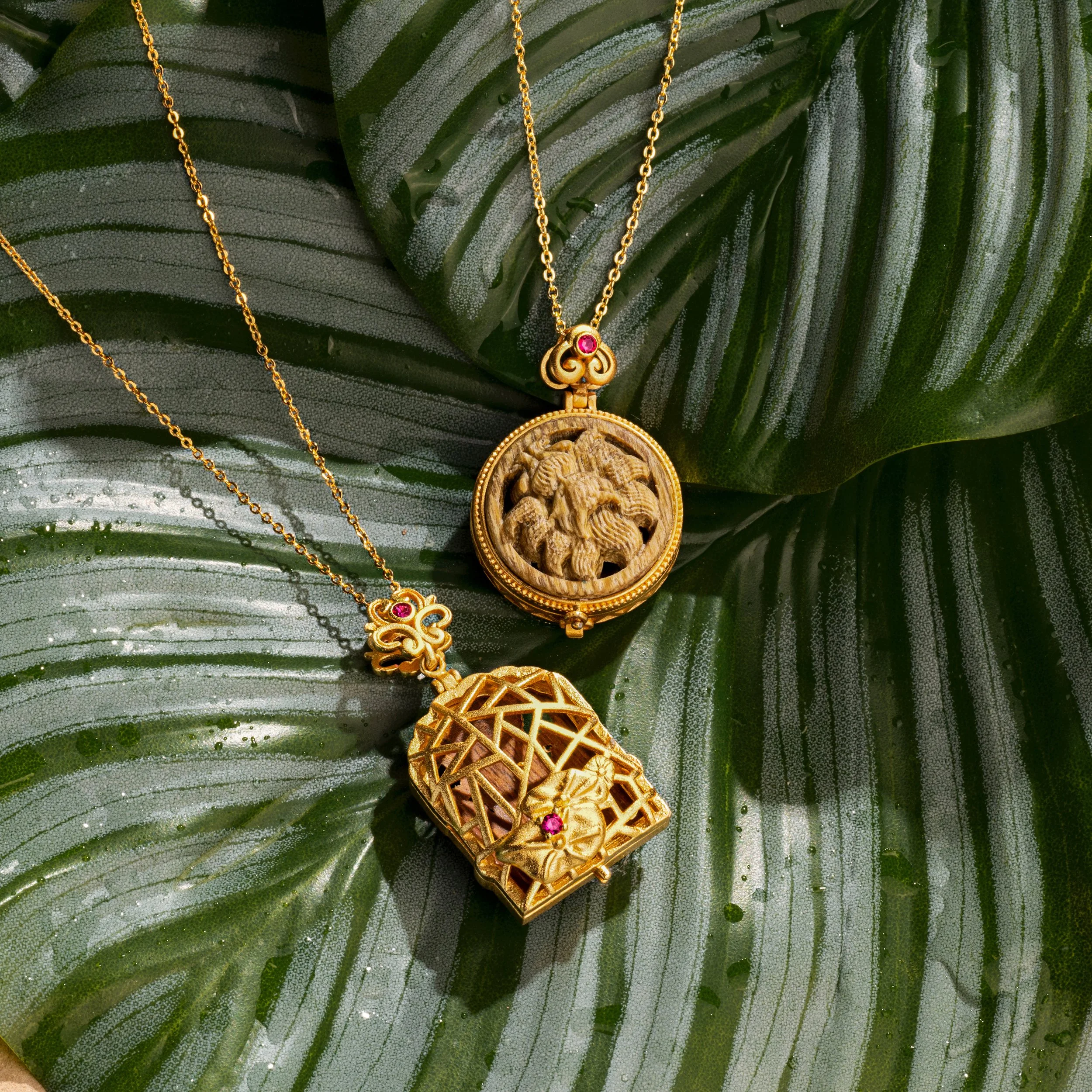 Two gold necklaces with ornate pendants resting on a green leaf. One pendant has a carved design, and the other has a lattice structure with a small gemstone inside.