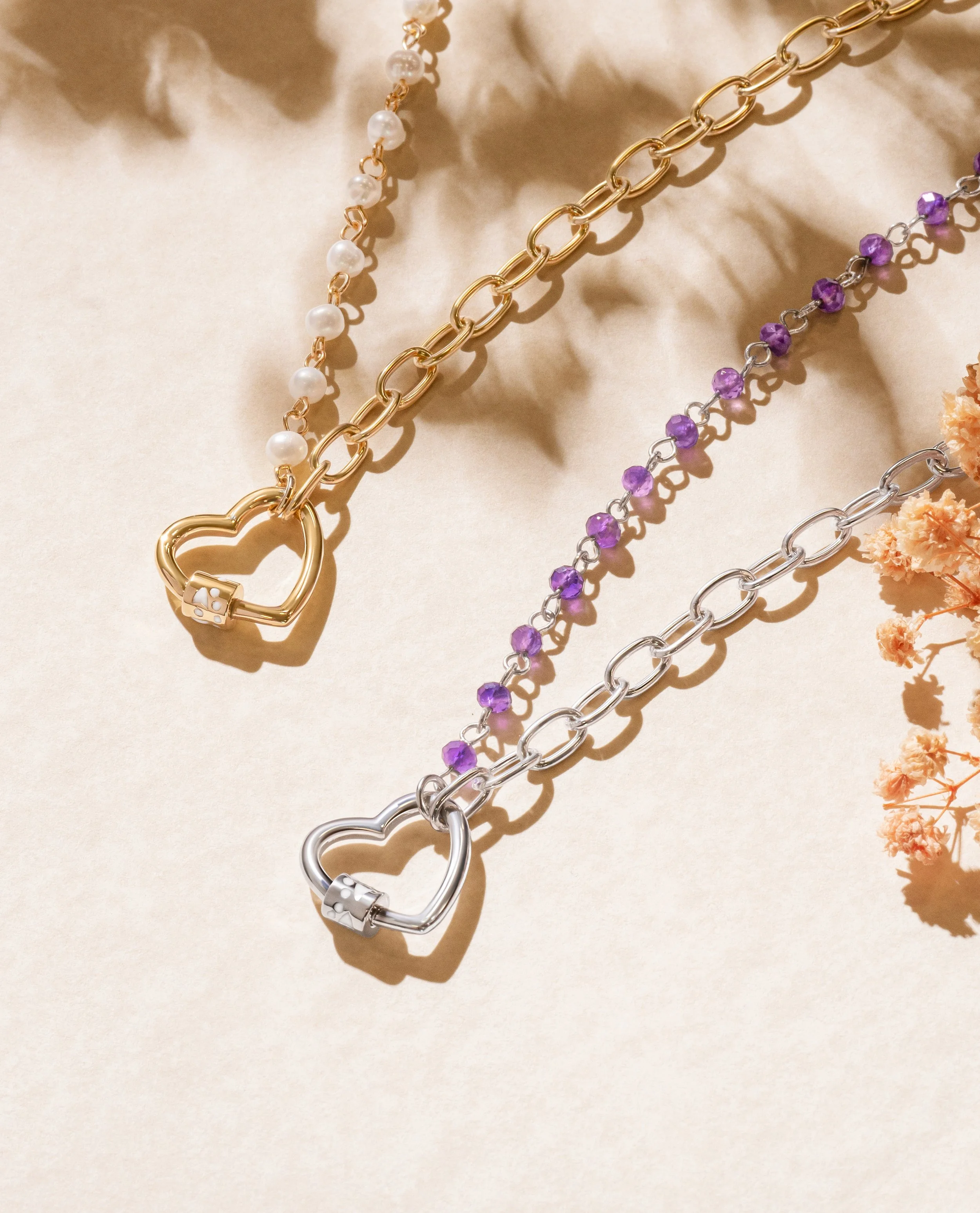 Three necklaces with heart-shaped lock pendants, one gold with pearl accents, one silver with purple beads, and one gold with silver chain links, arranged on a beige surface with dried flowers nearby.