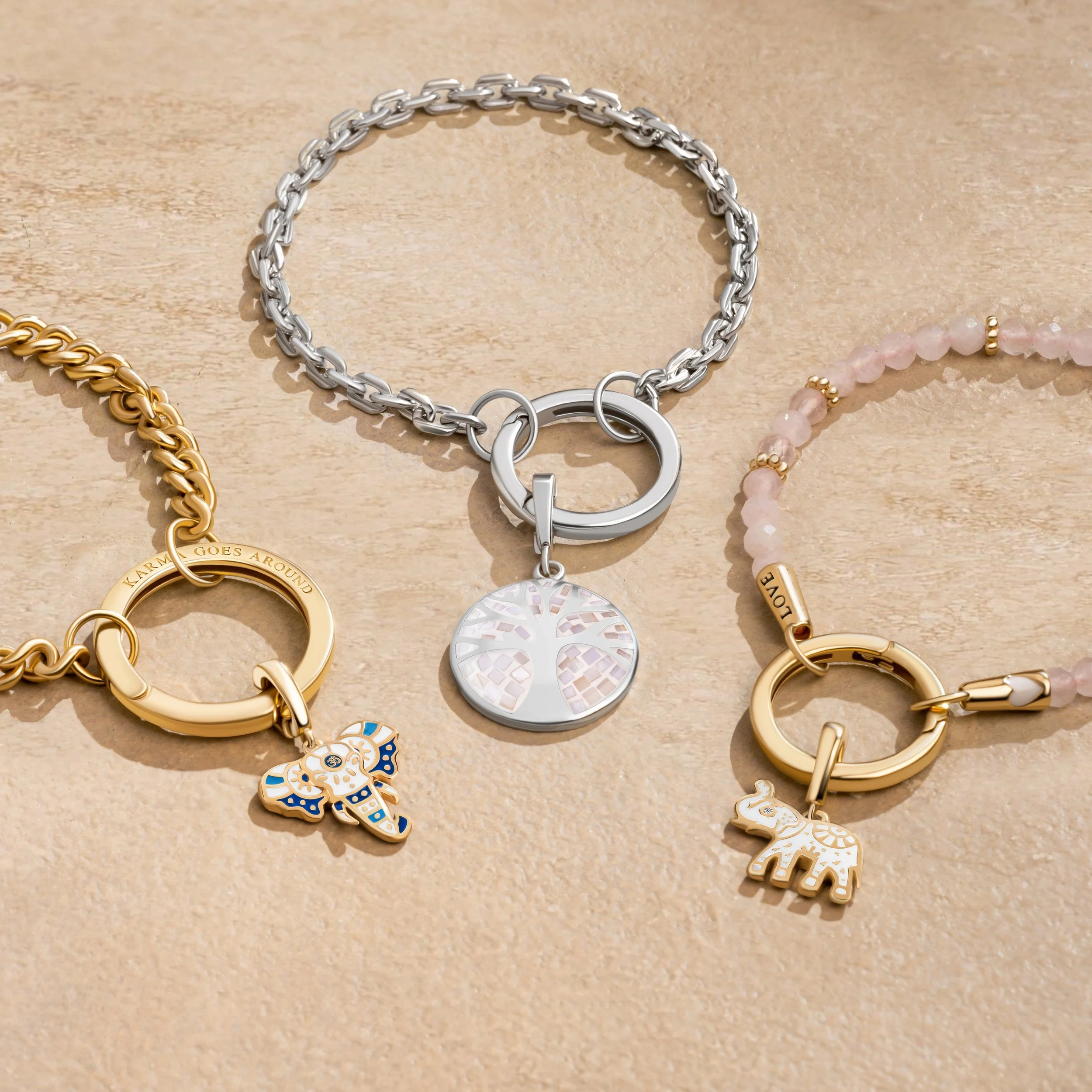 Three charms necklaces on a light wooden surface with silver, gold, and pink beads, featuring animal and tree designs.