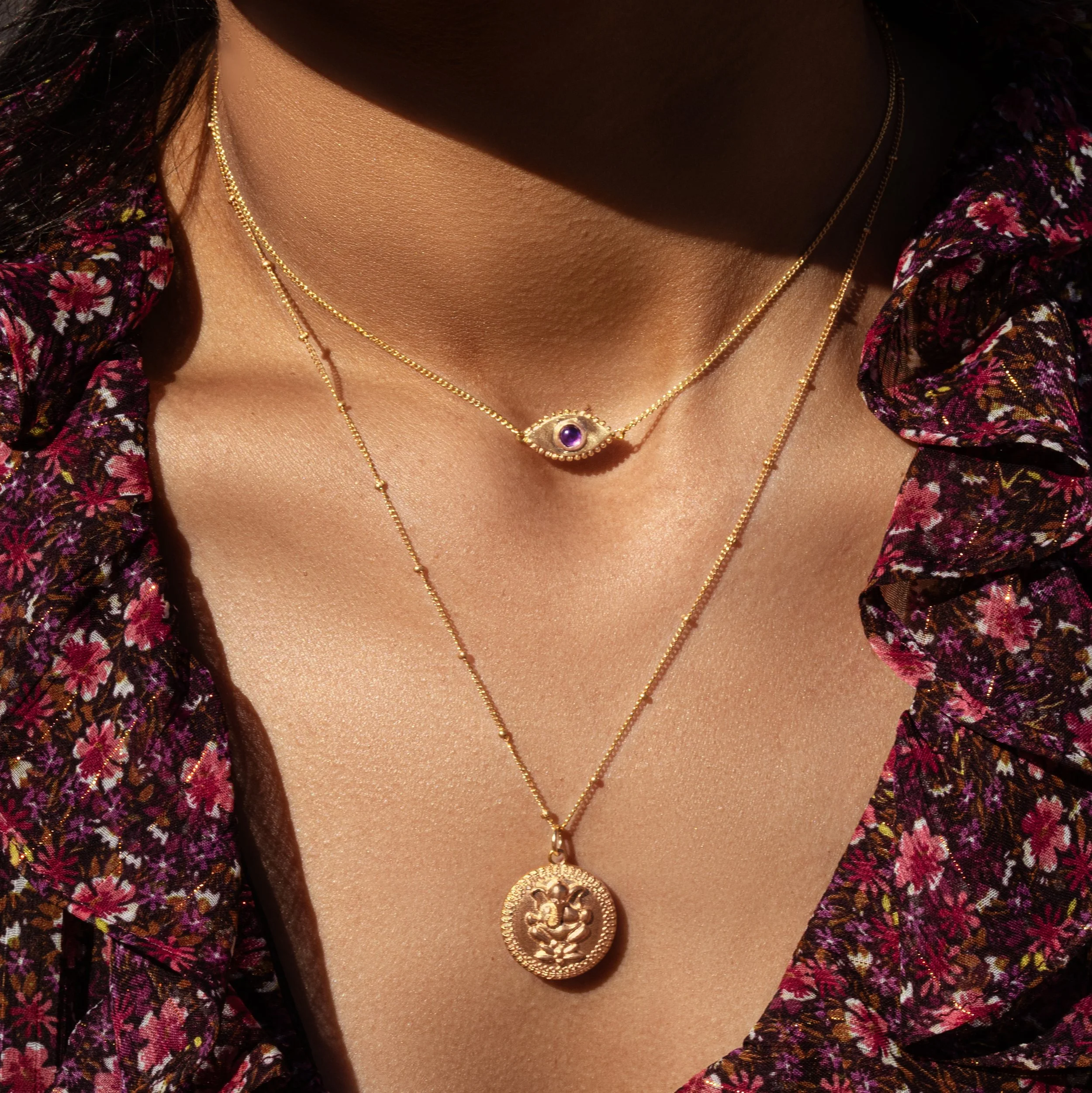 Close-up of a woman's neck and chest wearing layered gold necklaces, one with a purple eye-shaped pendant and another with a round medallion featuring a deity, against a floral patterned blouse.