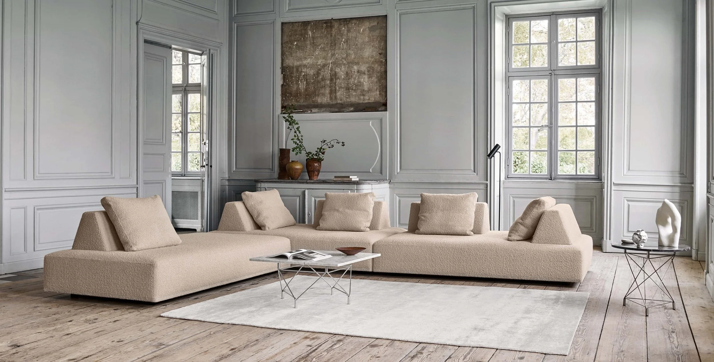 Modern beige sectional sofa with matching cushions in a spacious living room with wooden floors, gray paneled walls, large windows, a white area rug, and minimal decor including a coffee table, side table with sculptures, and a vase with green branches.