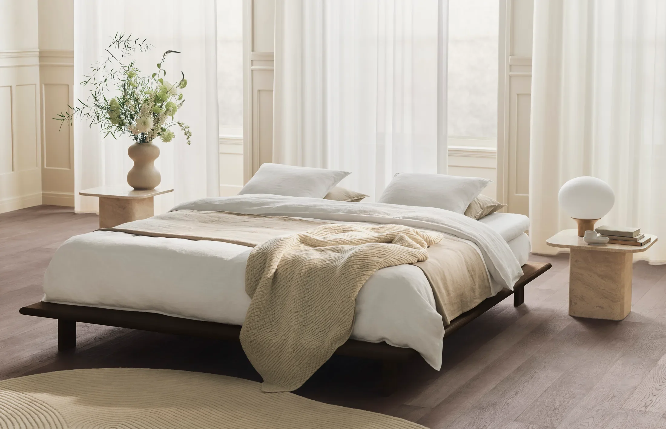 A cozy bedroom with a large bed dressed in white linens and beige blankets, flanked by two nightstands with modern lamps, and decorated with large windows with sheer white curtains.