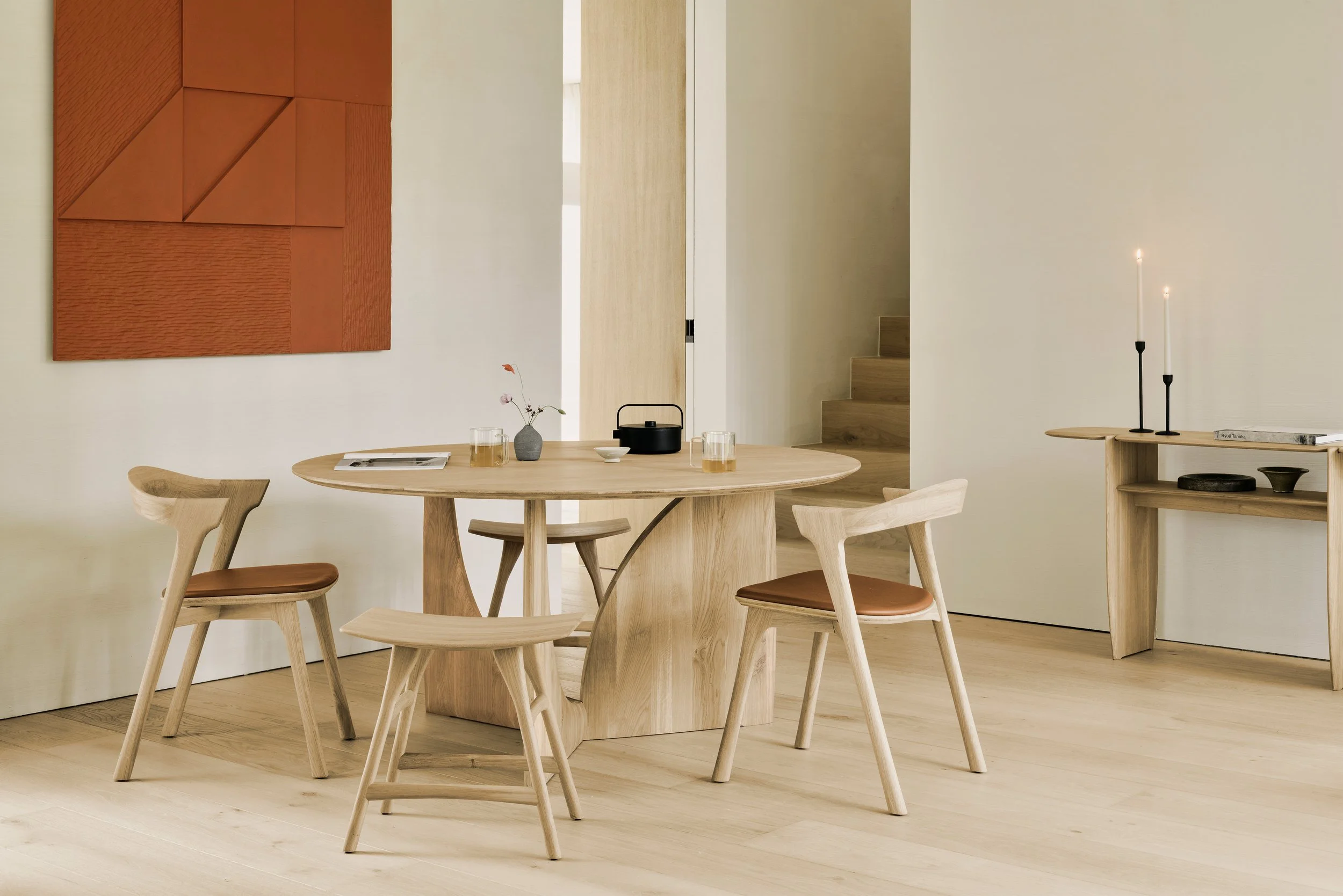 Minimalist dining area with a round wooden table, four matching wooden chairs with brown leather seats, a small side table with a candle holder and a book, and a wall-mounted abstract artwork in warm brown tones. The room has light-colored wooden flooring and walls, with stairs leading up in the background.