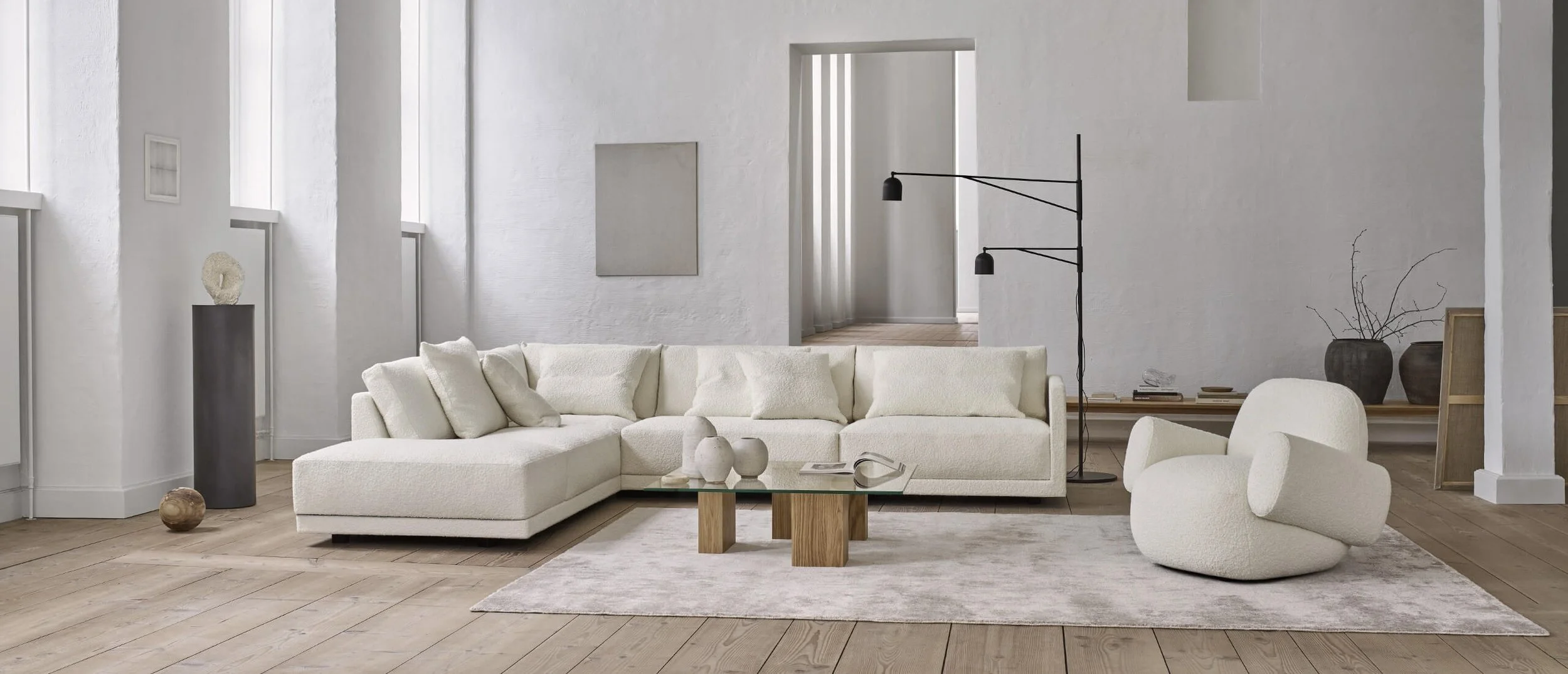 Minimalist living room with a white sectional sofa, a modern white armchair, a glass coffee table with wooden legs, a light-colored area rug, and black double-arm floor lamps, all set in a bright space with white walls and light wood flooring.