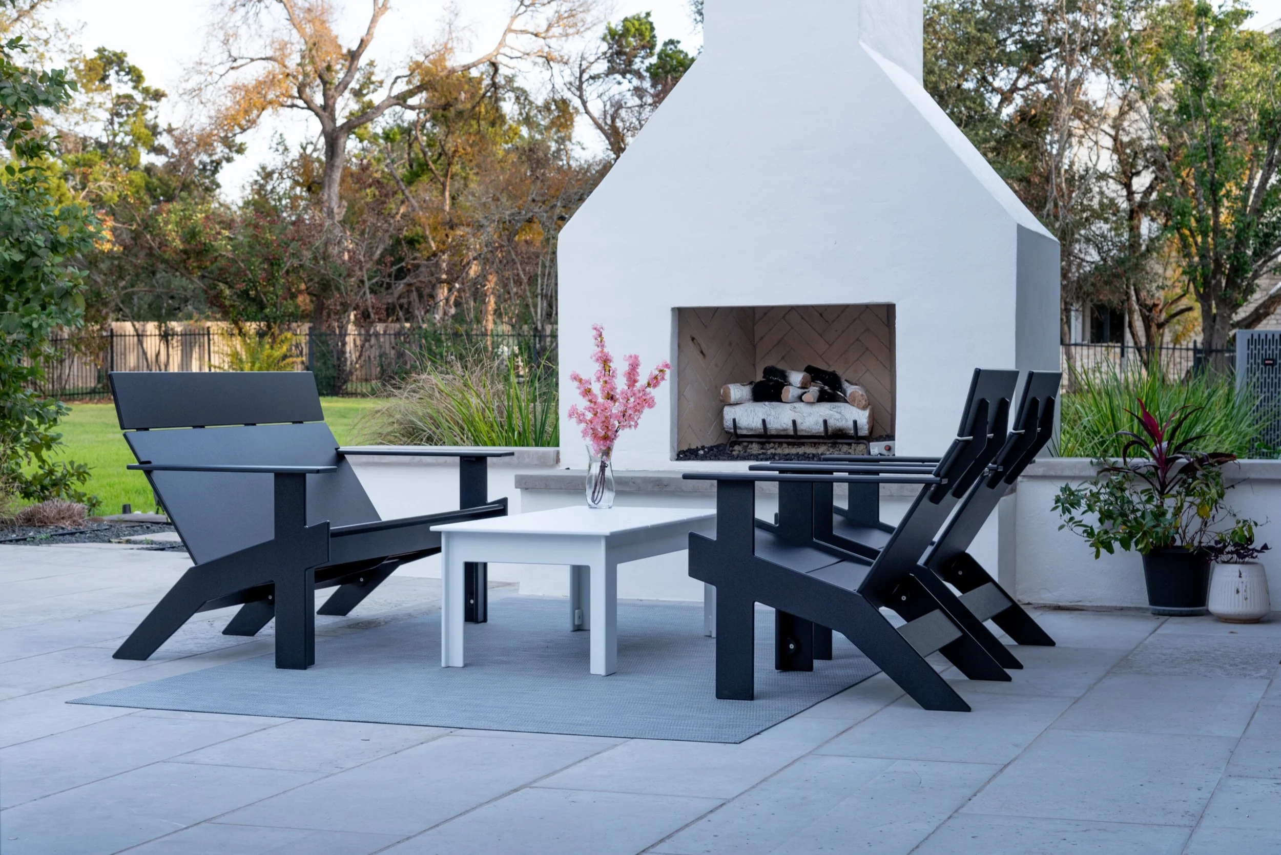 Outdoor patio with black Adirondack chairs around a white table, pink flowers in a clear glass vase, potted plants, and a white outdoor fireplace with logs inside, on a tiled patio surrounded by trees and greenery.