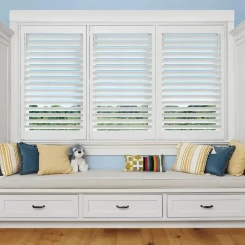 A window with white plantation shutters in a light blue wall room, with a built-in white seating area below, decorated with yellow and blue cushions and a stuffed dog toy.