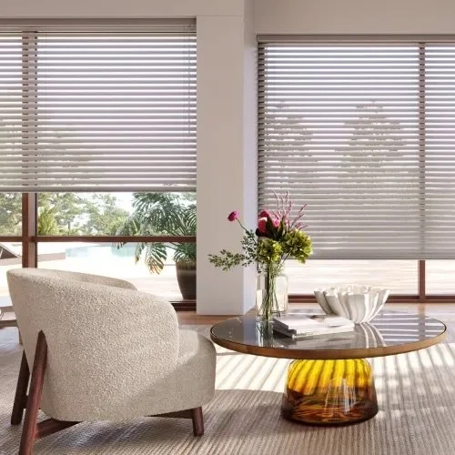 Living room with large windows, a beige armchair, a glass-topped coffee table with a yellow glass base, a tall vase with flowers, and a decorative white bowl.
