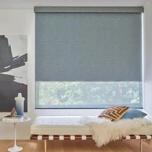 Modern bedroom with large window covered by a grey roller shade, a white chaise lounge with pillows and a white furry throw blanket, and a side table with decorative items.