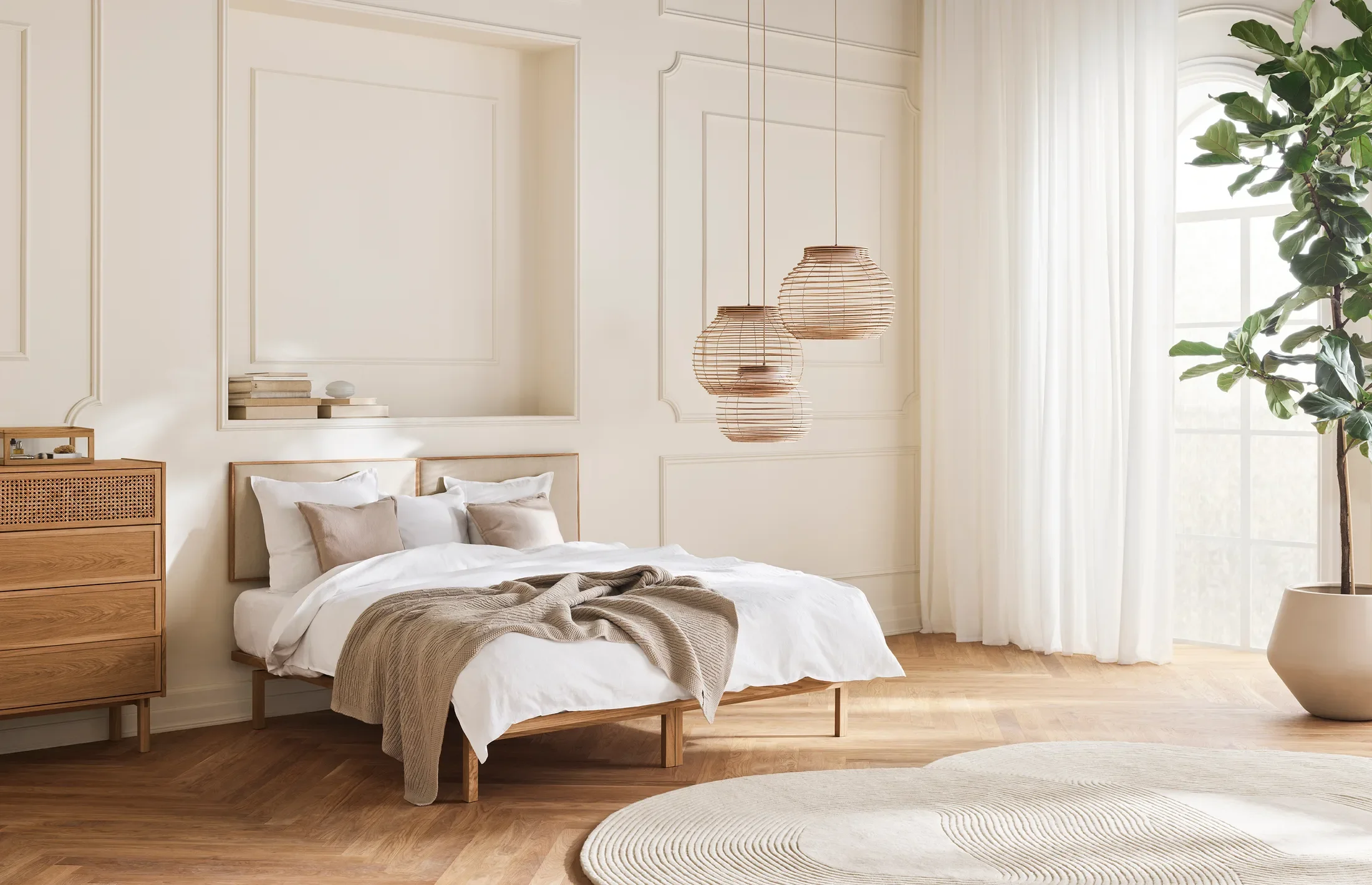 A bedroom with a large bed, beige pillows, a beige blanket, wooden flooring, a wooden dresser, hanging pendant lights, white curtains, and a potted plant near a large window.