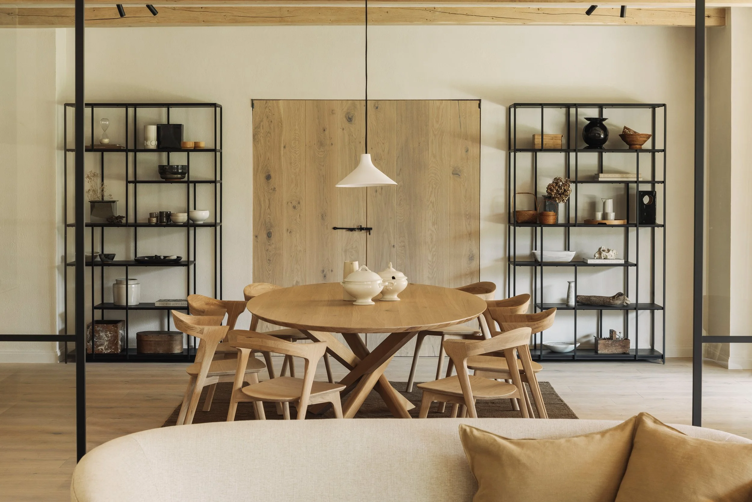 A modern dining room with a wooden round table, six wooden chairs, black metal shelves filled with decorative items, a white pendant light, and a beige sofa with a gold pillow visible in the foreground.