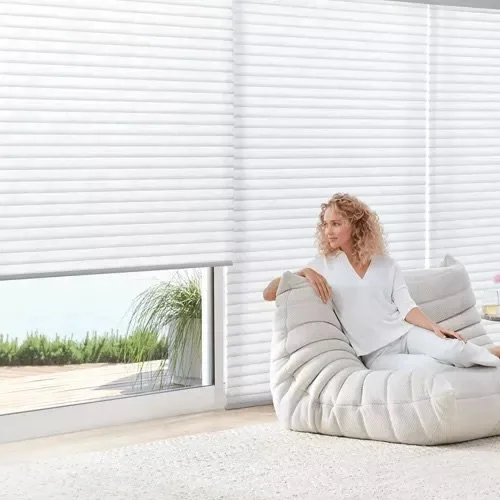 A woman sitting on a modern, white, tufted sofa in a bright room with large window blinds and a view of a garden outside.