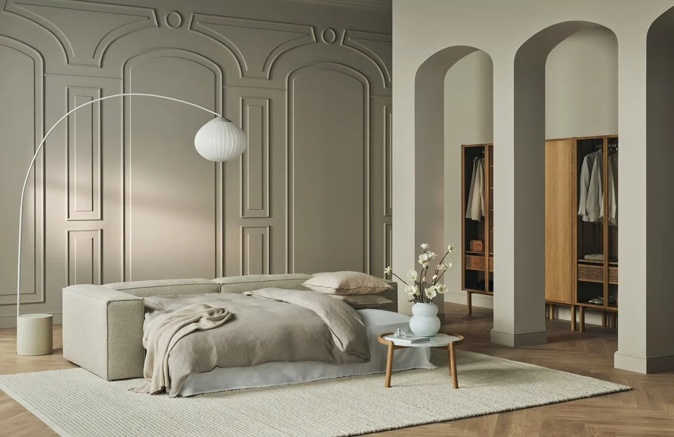 A modern bedroom with neutral tones, a beige upholstered bed with linen bedding, a small wooden side table with a white vase containing white flowers, and built-in wooden wardrobes, with an arched entryway and decorative wall paneling.