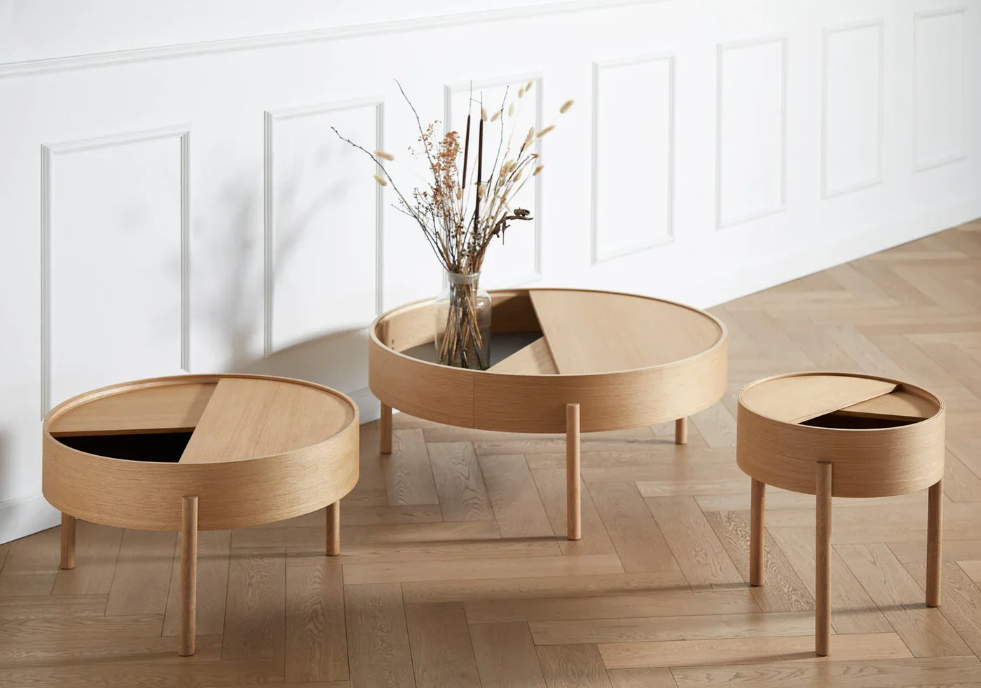 Three round wooden tables with sliding tops, one with a glass vase containing dried flowers, set against a white paneled wall with hardwood flooring.