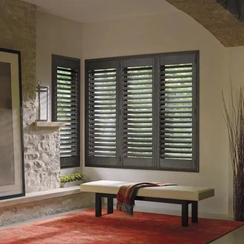 A room with large gray window shutters, a stone fireplace, a white bench with a blanket, and a red rug.