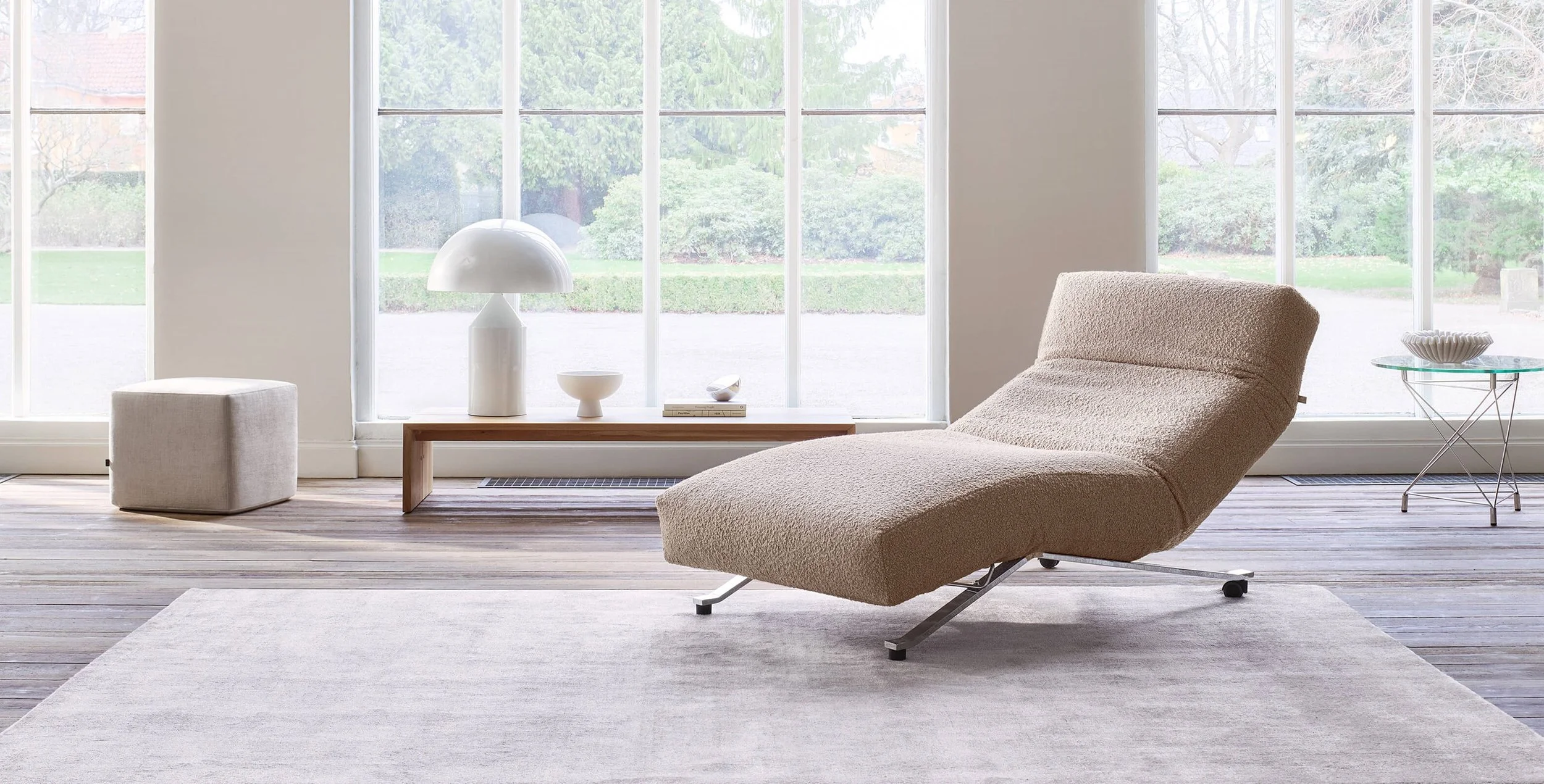Modern living room with large windows, beige chaise lounge, white side table, white ottoman, and decorative lamps, overlooking a green outdoor space.
