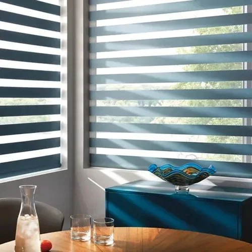 Sunlight streams through horizontal blinds into a modern room with a blue cabinet, a decorative glass bowl, and a table with a pitcher and glasses.