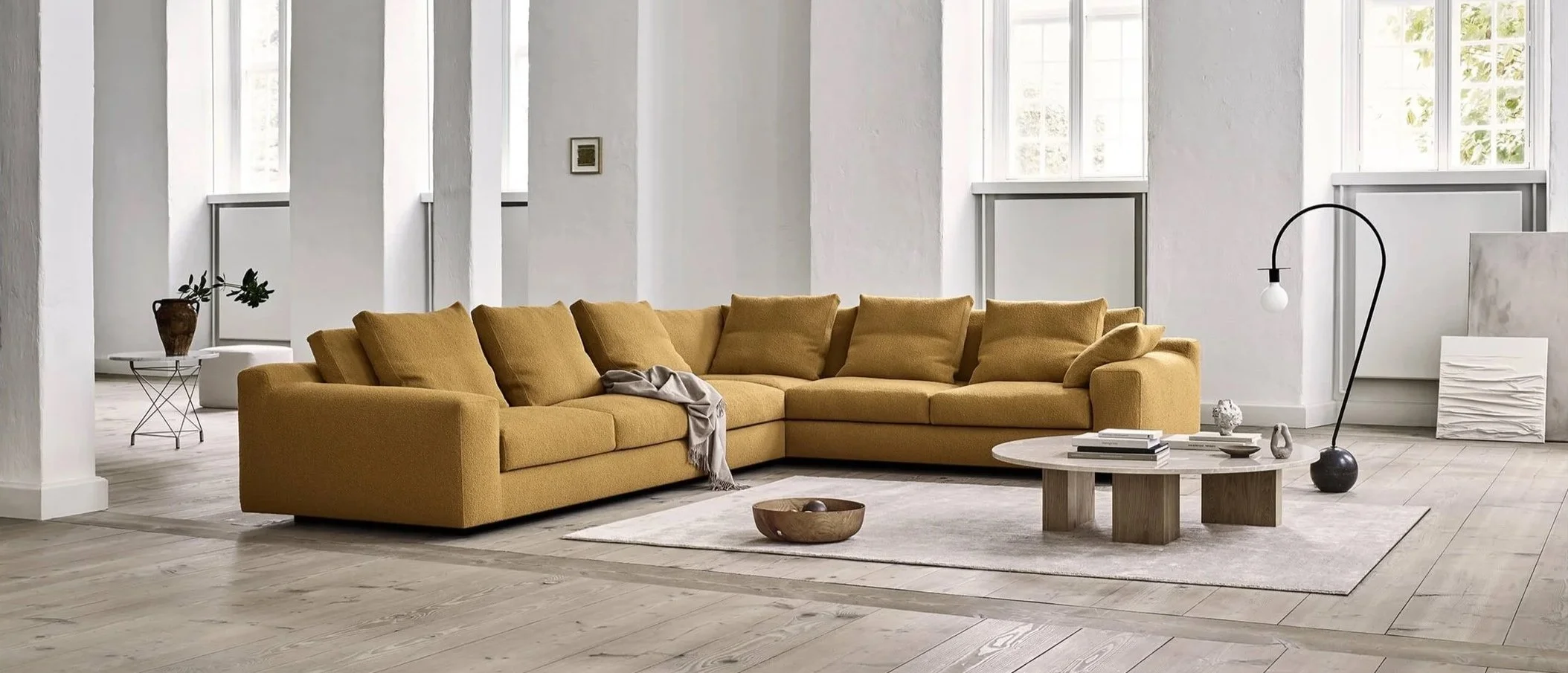 Living room with a large mustard yellow sectional sofa, a round wooden coffee table, and minimalist decor including a floor lamp, books, and abstract art in a bright space with white walls and windows.