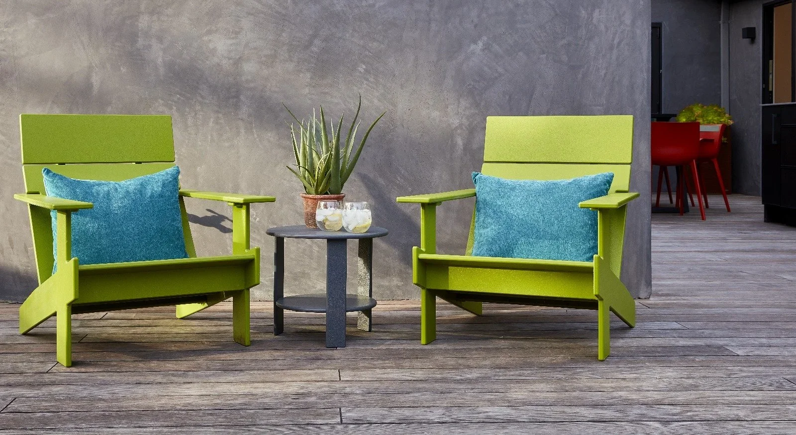 Two lime green outdoor chairs with blue pillows, a small black round table with a potted plant and two glasses of drinks, and a wooden deck floor, with a gray wall in the background.