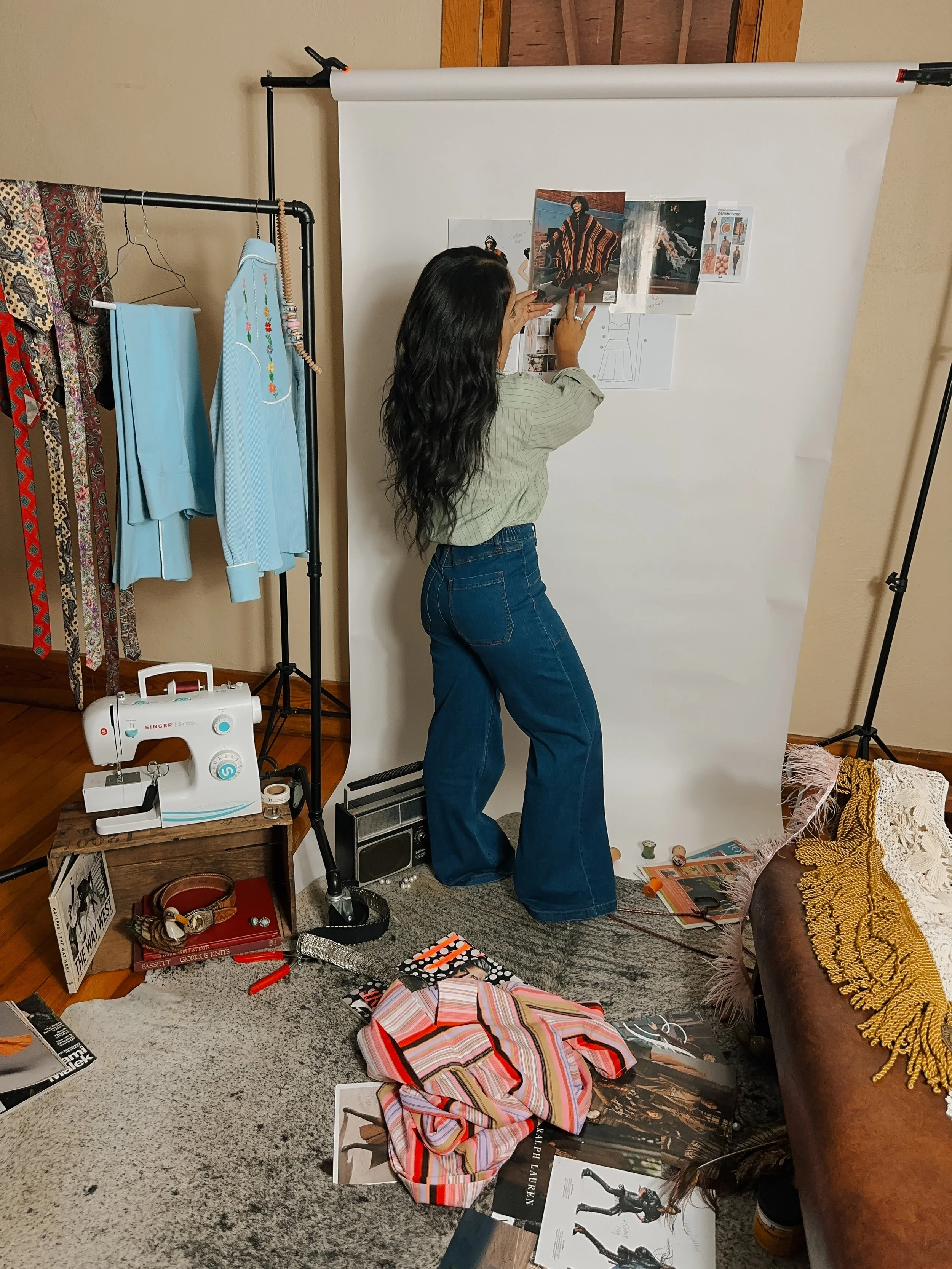 A woman in a striped shirt and blue jeans arranging photographs and mood boards on a white backdrop in a room. The room has clothing on a rack to her left, a vintage sewing machine on a wooden crate, and various art and fashion-related items on the floor and furniture.