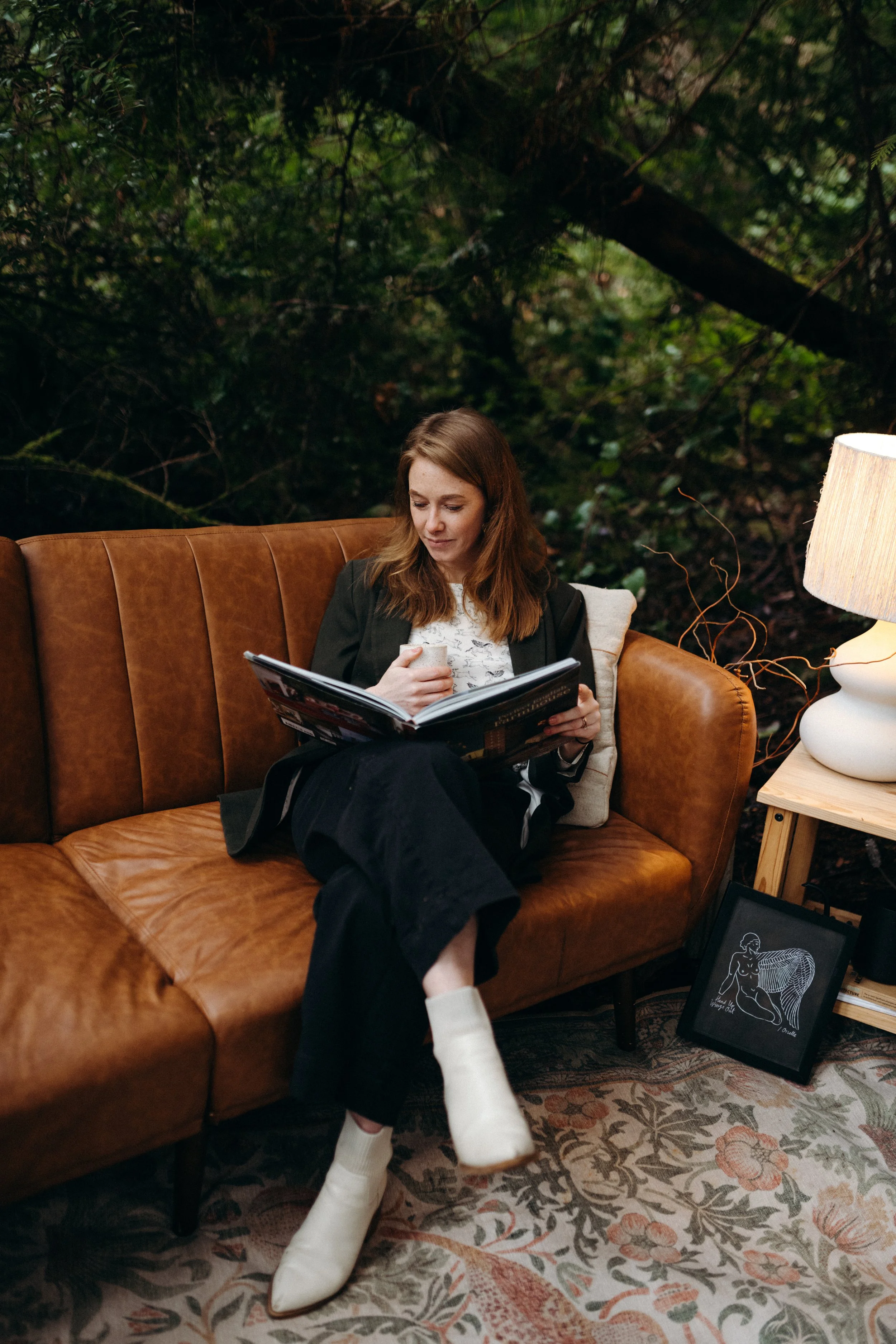 A woman is sitting on a brown leather couch, reading a magazine and holding a drink. She is in a cozy indoor space with a painting on the floor and a lamp on a side table. Outside the window behind her, there are trees and greenery.
