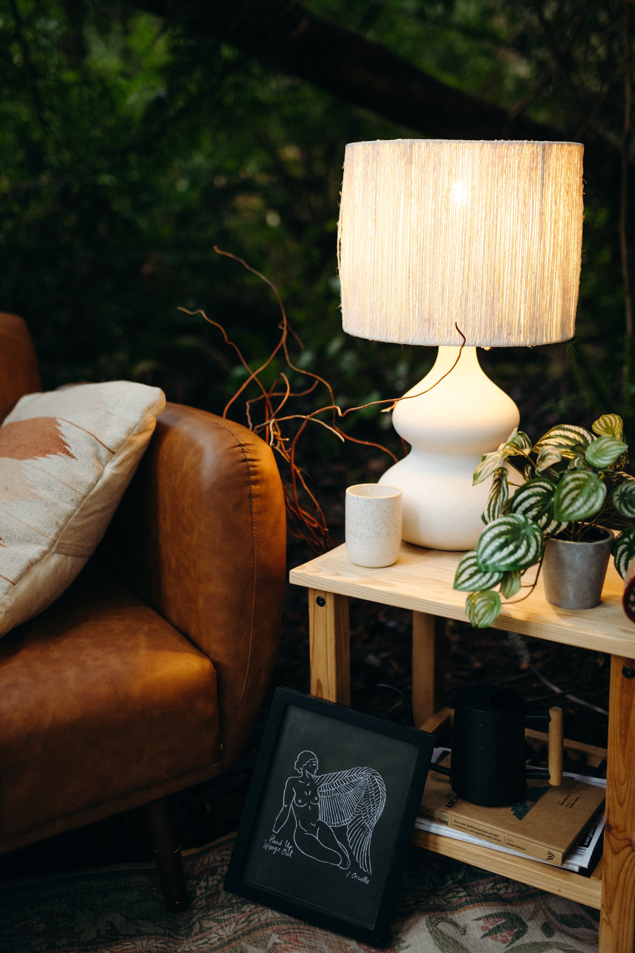 A cozy living room corner with a leather sofa, a wooden side table holding a large white lamp and potted plants, and a framed art piece leaning against the sofa.