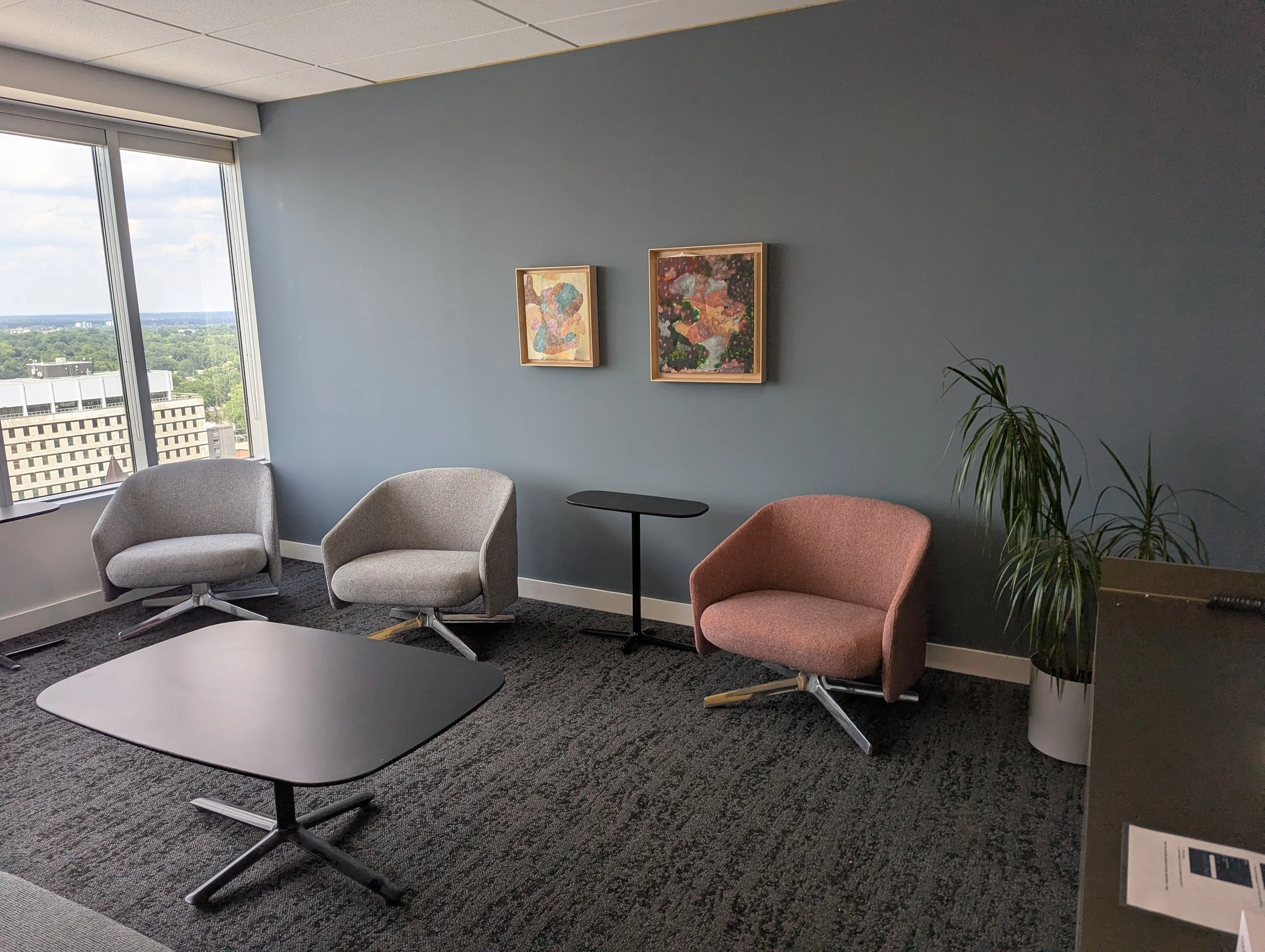 A modern office waiting area with two gray and one pink accent chairs, large windows showing a cityscape with trees, two framed abstract paintings on a blue wall, a small black side table, and a green plant in a white pot.