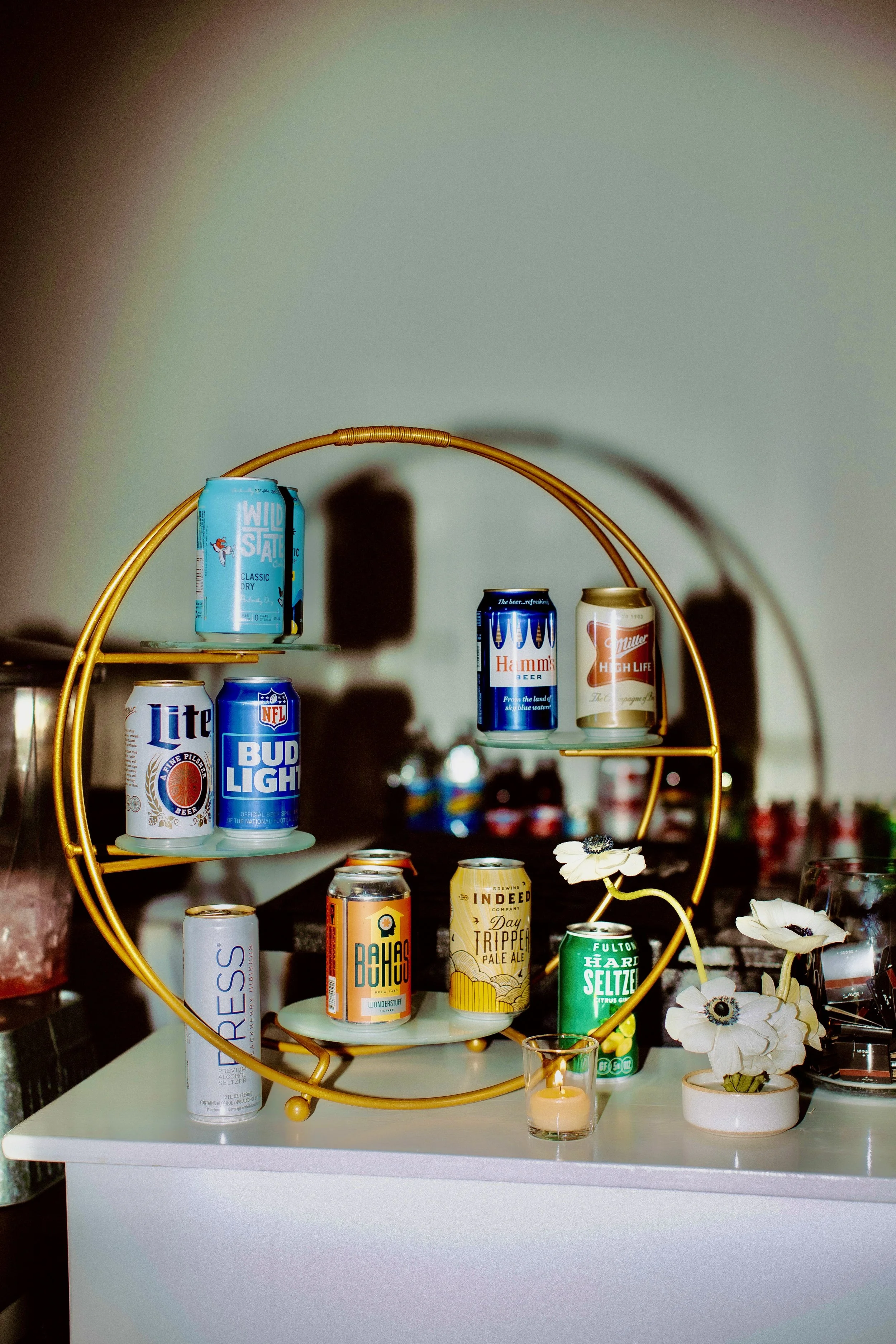 An assortment of various beer and beverage cans displayed on a gold circular metal stand, set on a white surface. The stand holds different cans with distinct colorful designs. Nearby is a small candle in a glass holder and a decorative floral arrangement.