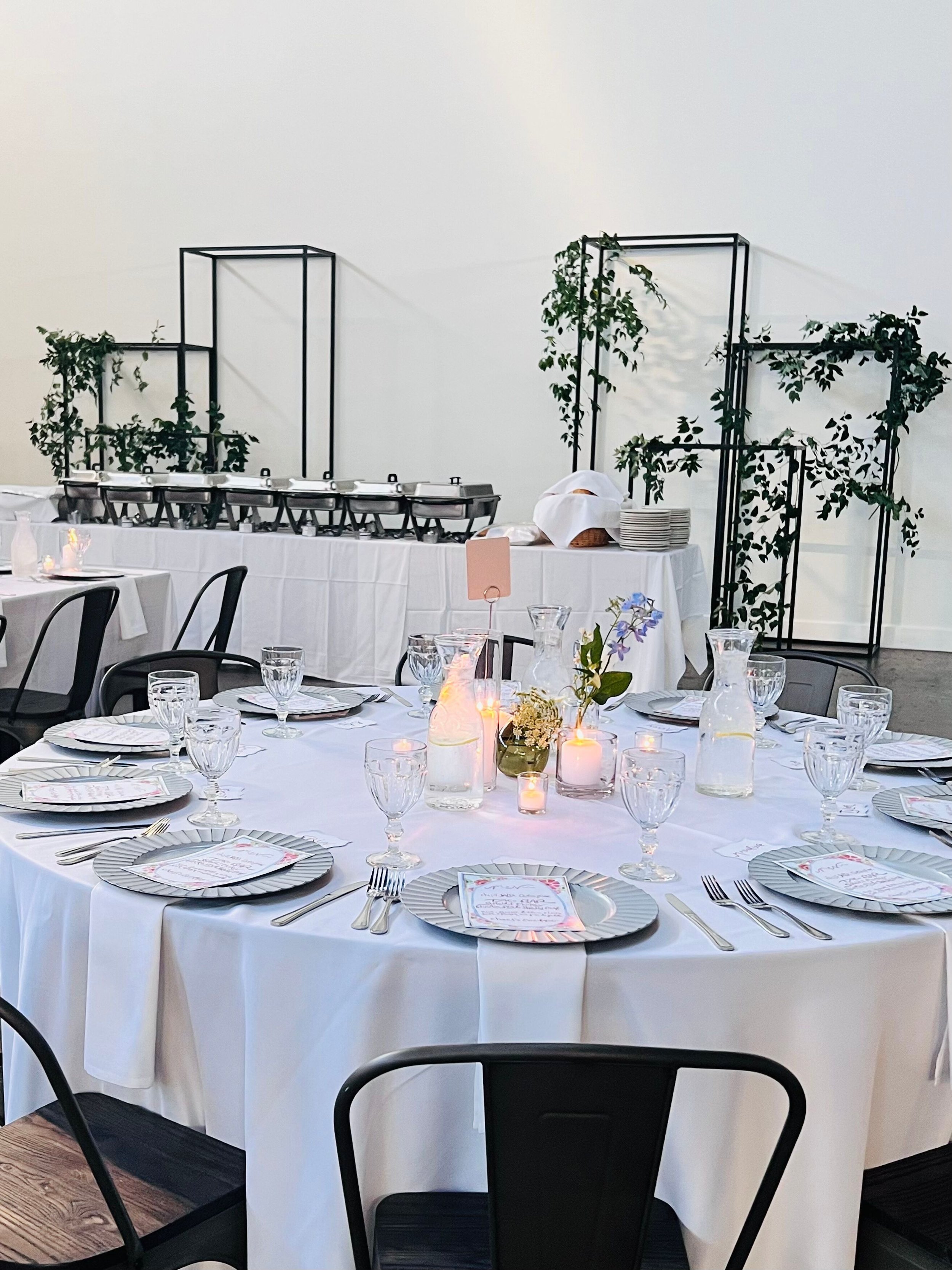 Dinner tables setting with bud vases, candles, charges, and buffet line in the background with chafers and black frames with greenery.