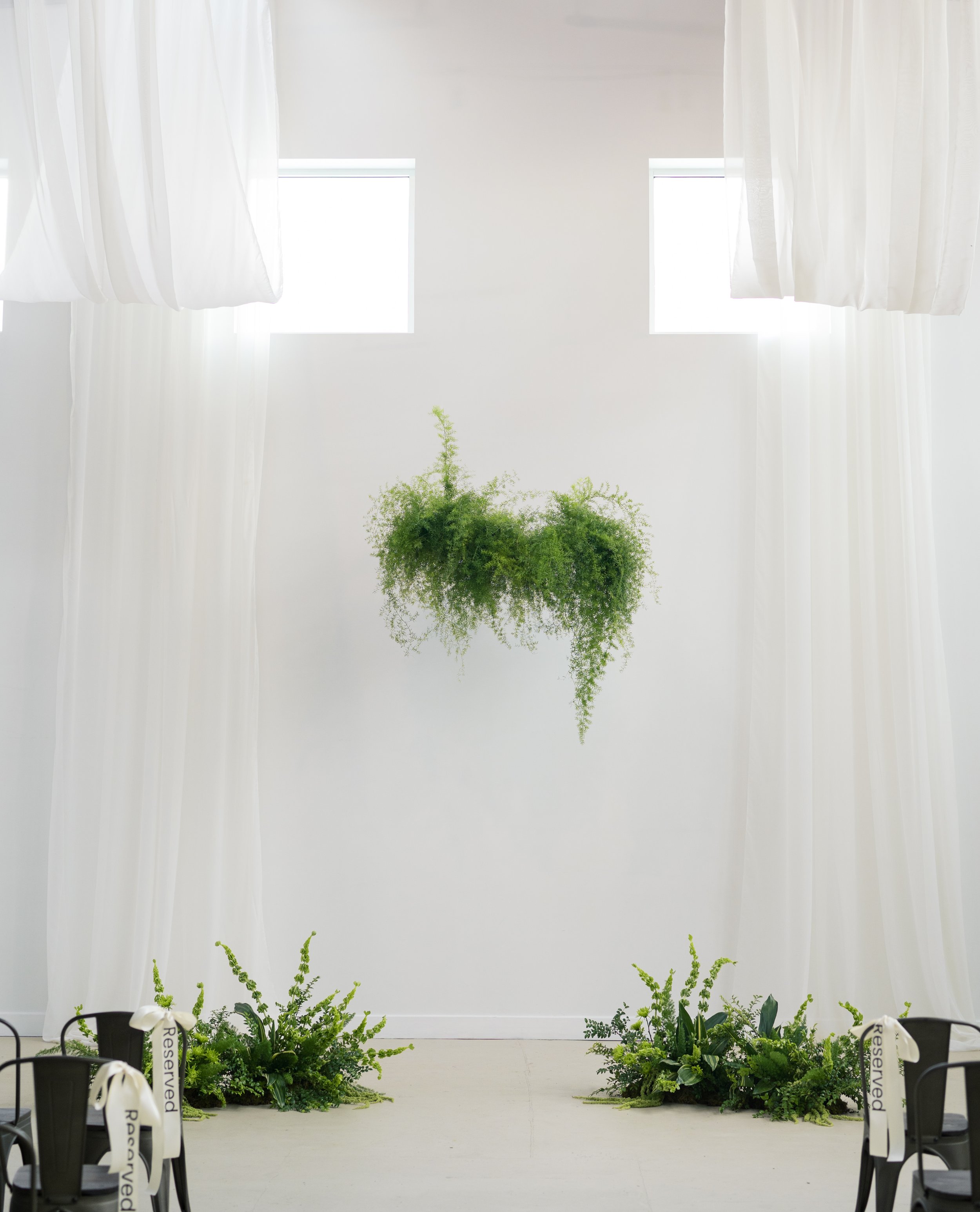 Pure white draping along a tall wall with windows, that includes a hanging fern and greenery sway, with 2 large floor arrangements to match.