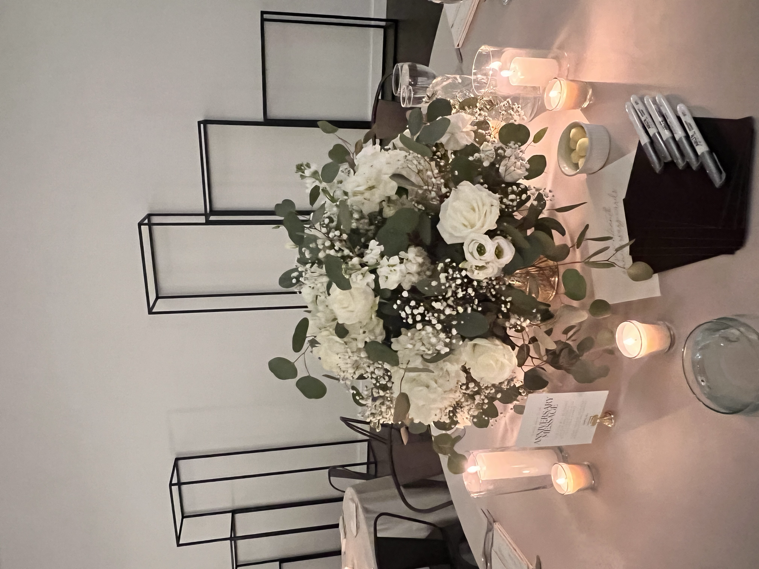 Large white and green table floral arrangement surrounded by different size candles, with black frames in the background to get dimension to the wall space.