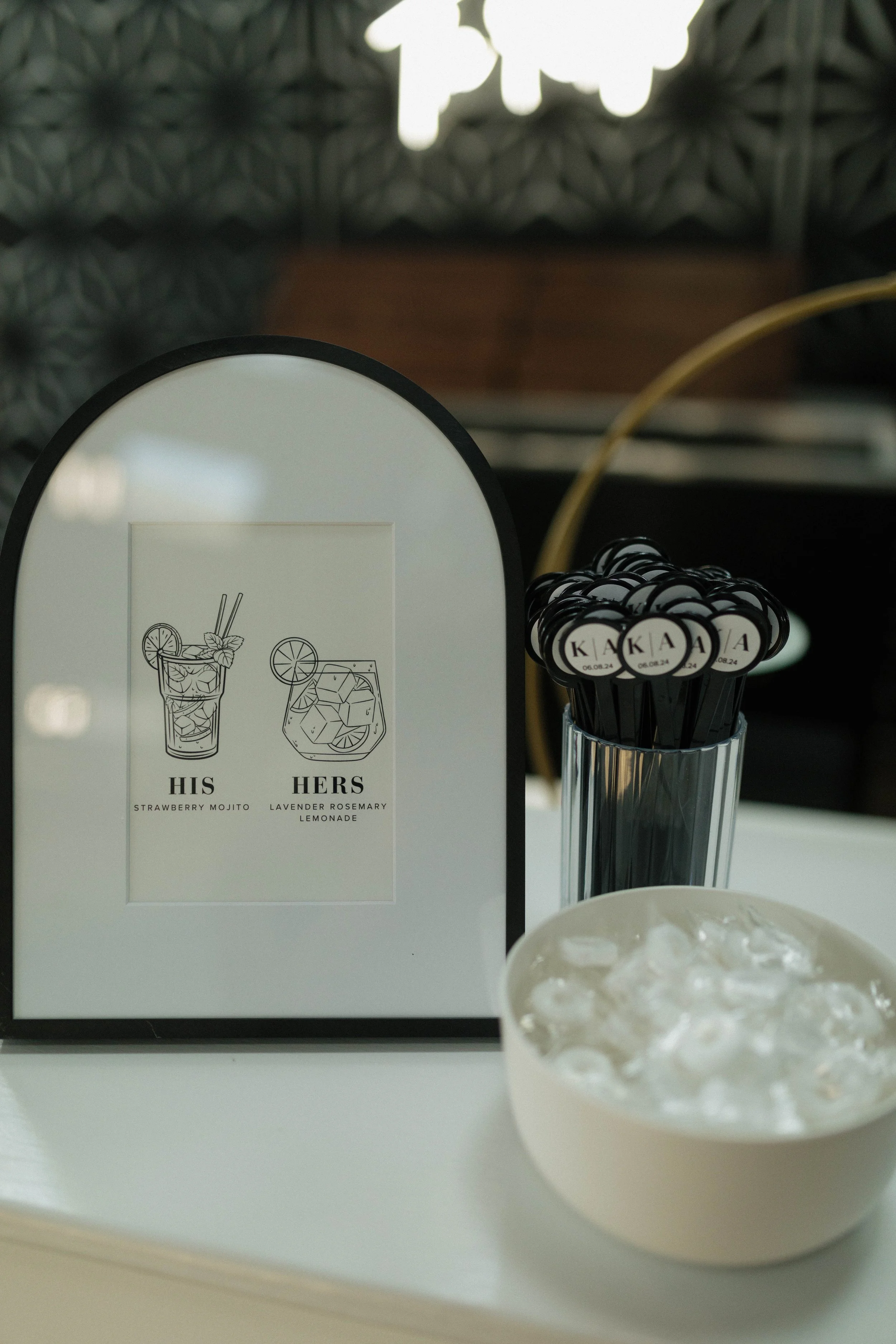 Framed cocktail menu with "His Strawberry Mojito" and "Hers Lavender Rosemary Lemonade," next to a cup with cocktail stirrers and a bowl of ice.