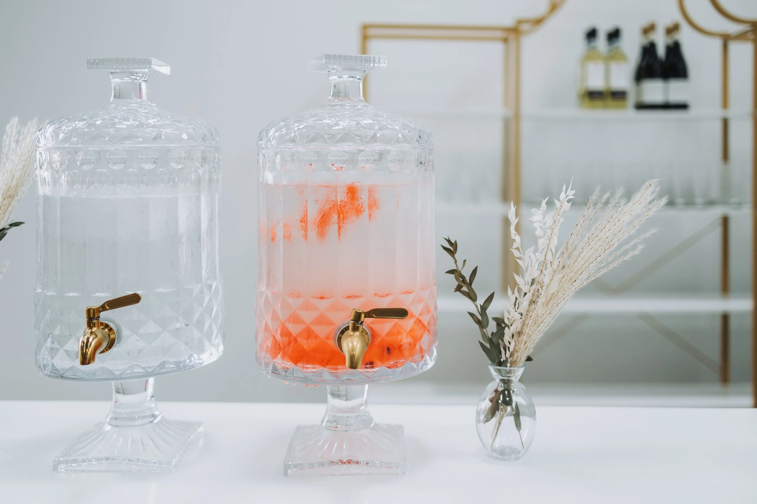 Two glass beverage dispensers with gold spigots, one with clear liquid and the other with pink liquid, on a table with a small vase of dried flowers.