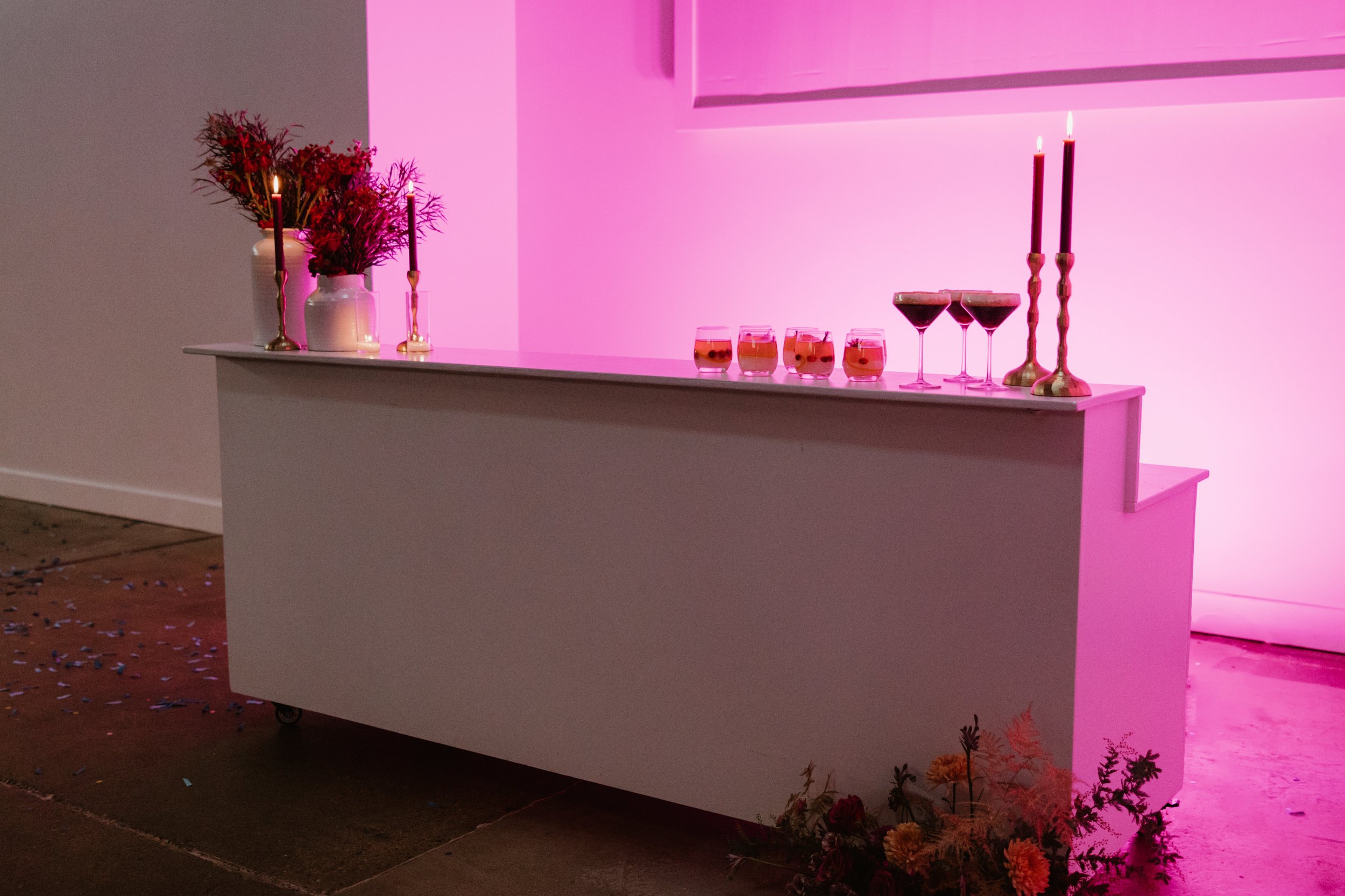 Decorative bar setup with pink lighting, featuring flowers in vases, lit candles, and various drinks on a white countertop.