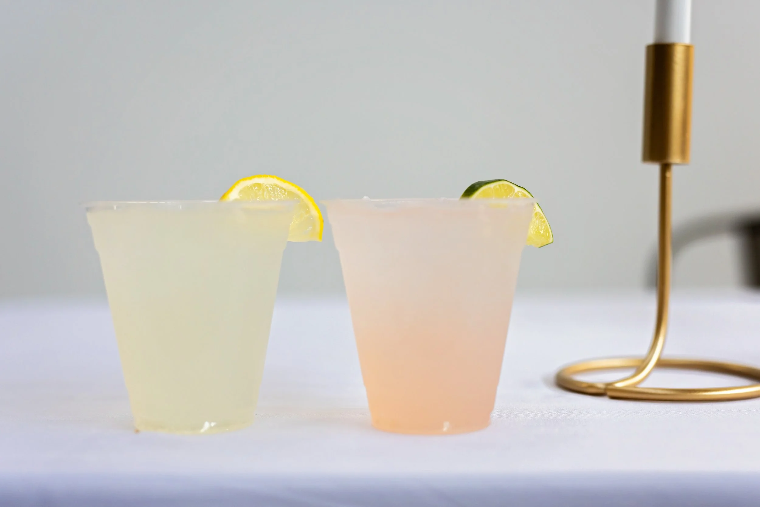 Two drinks in clear cups on a table, one with a lemon slice and the other with a lime slice garnish, next to a gold candlestick.