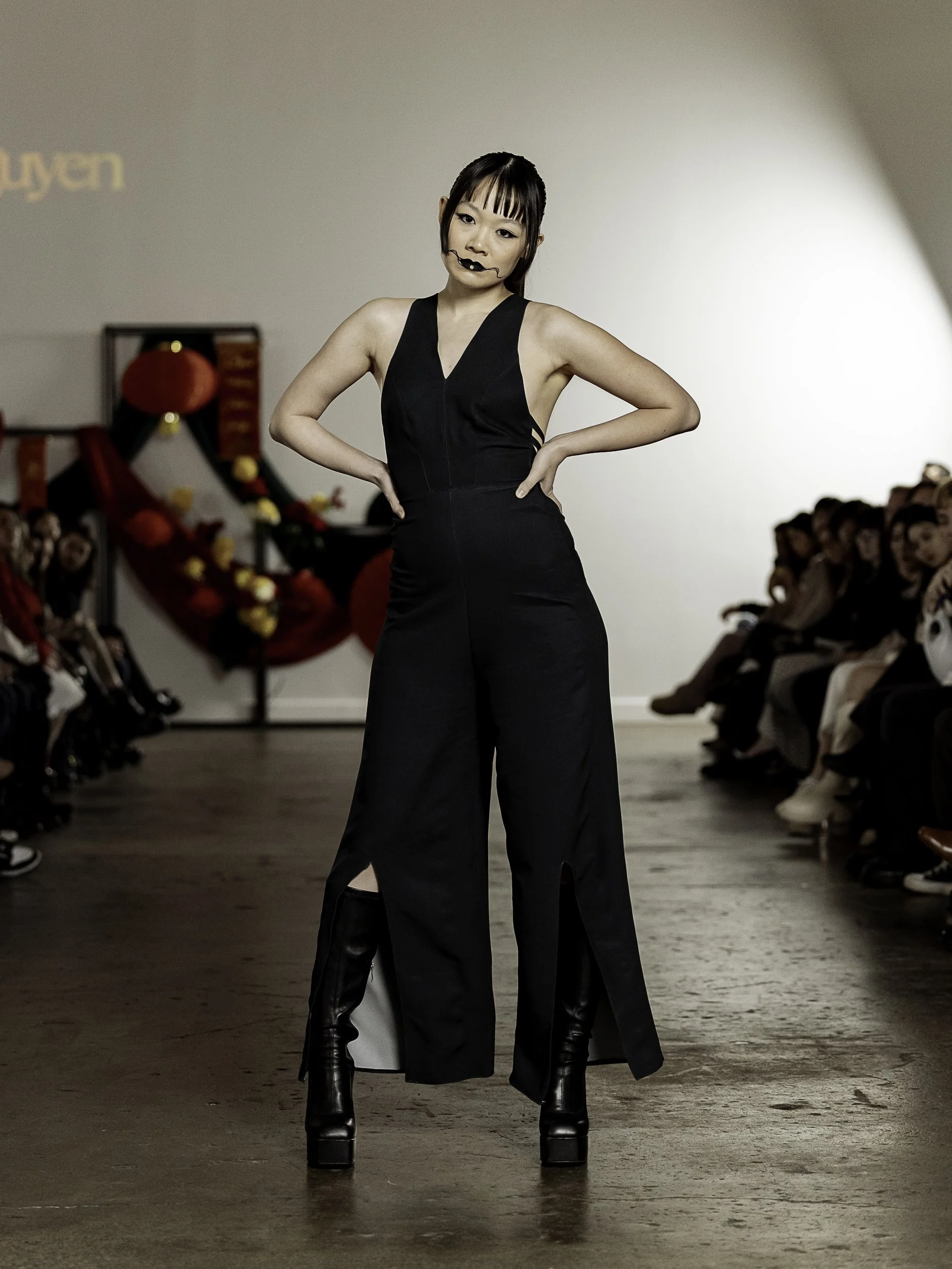Model in black jumpsuit with arms on hips posing for the camera during a fashion show at The Luminare
