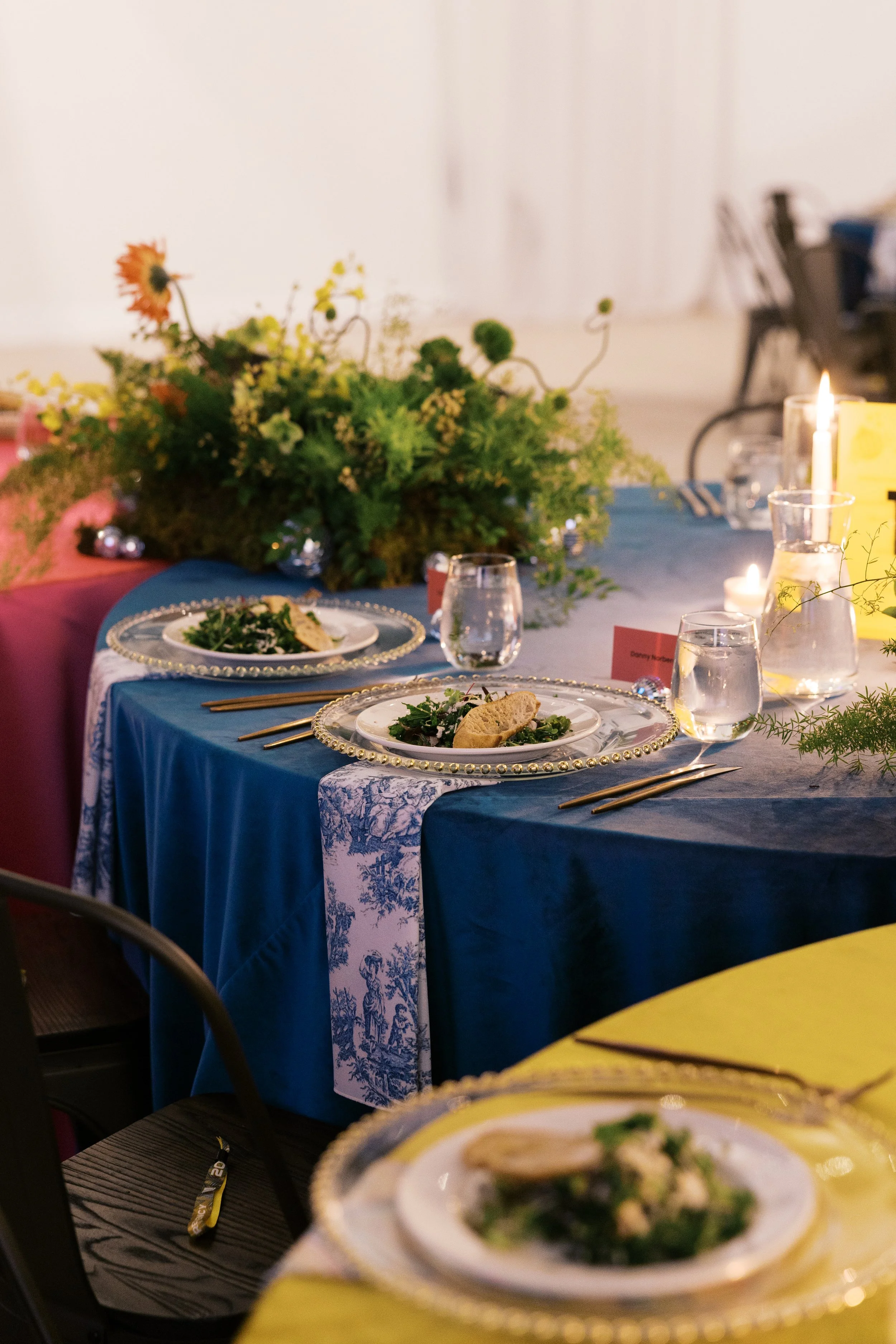 Dinner table setup with multiple colored linens in a caterpillar style layout, with preset salad, water service, candles.