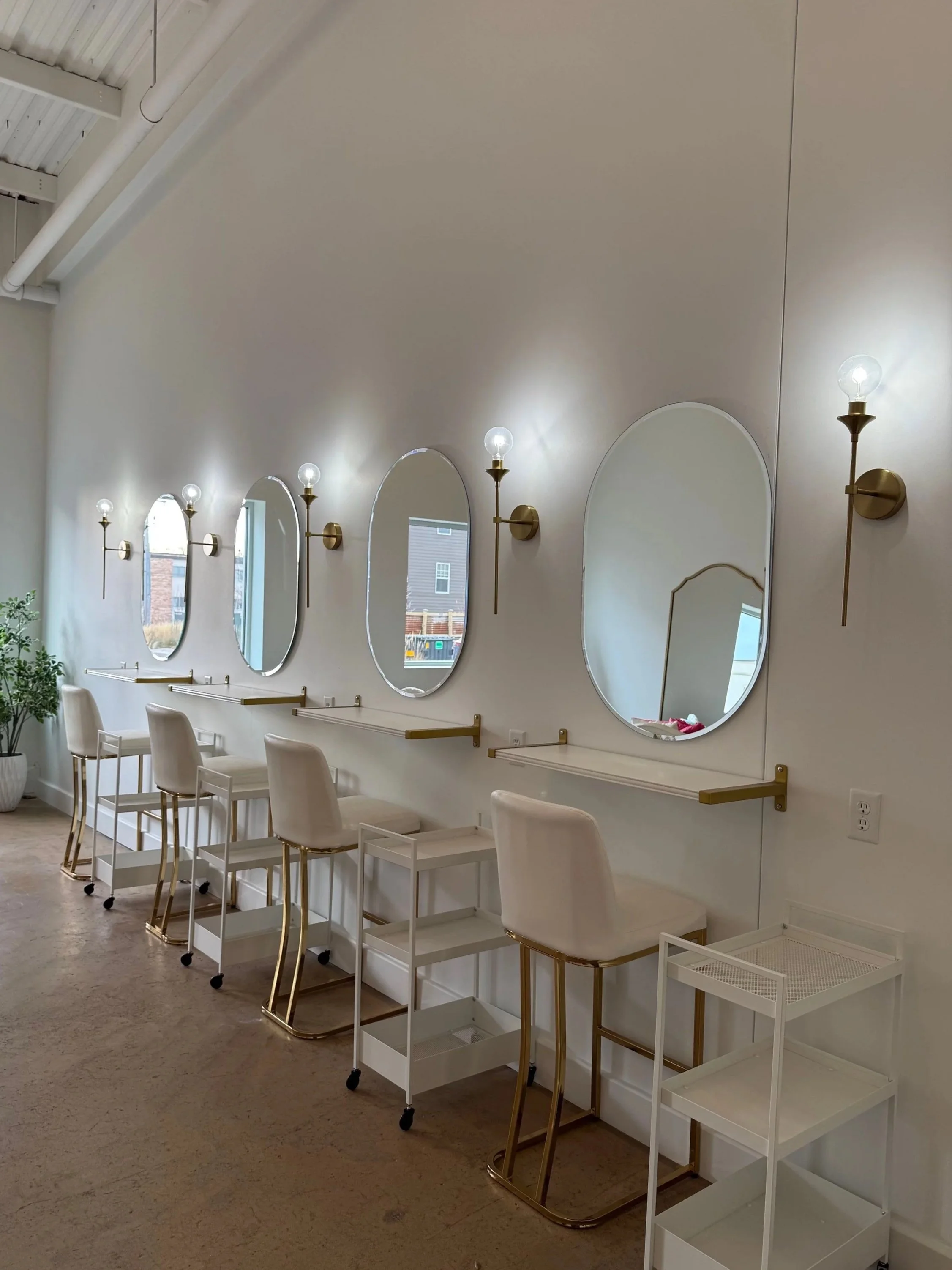 Makeup mirrors on a white wall with sconce lighting, in gold trim, with bar height white barstools.