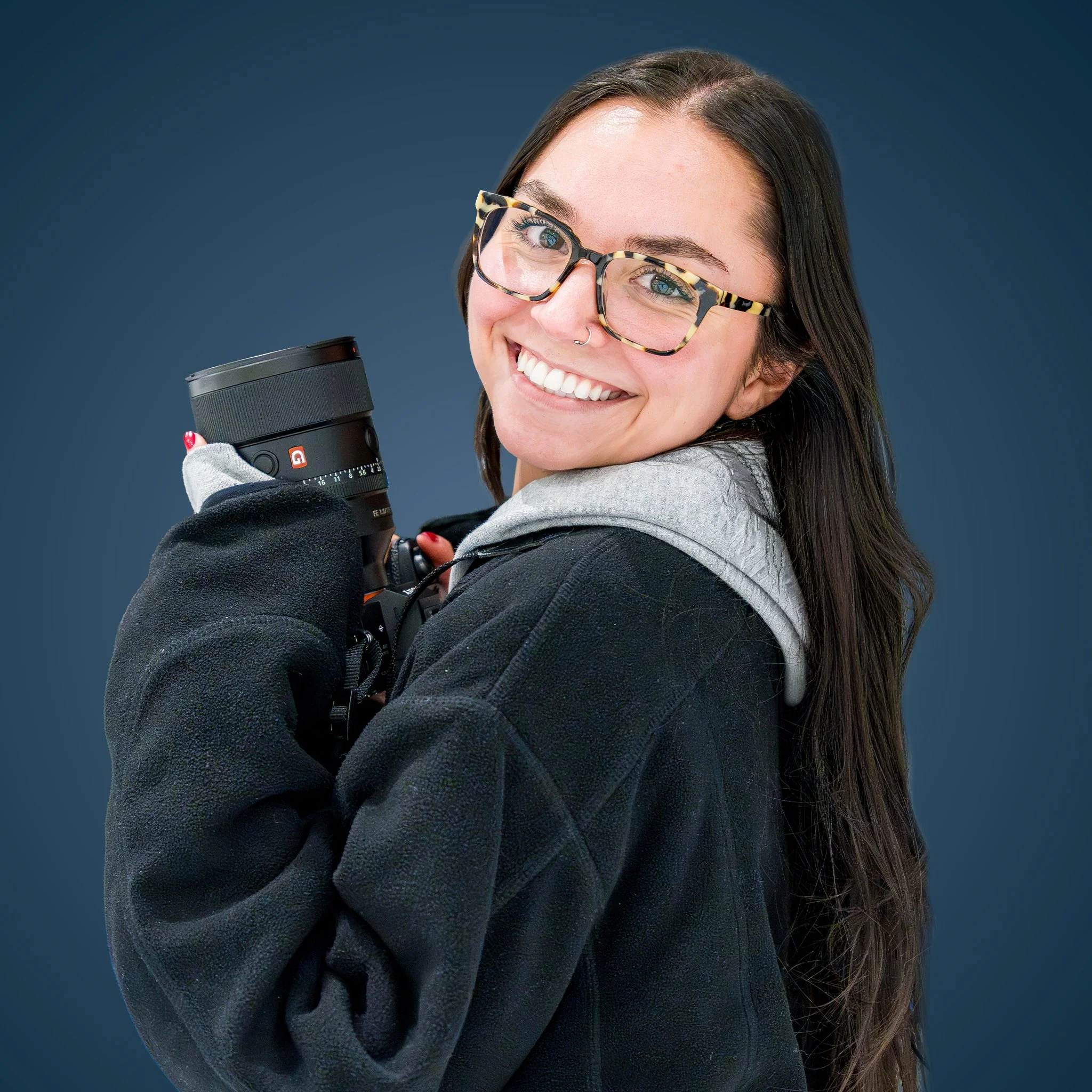 A young woman with long brown hair and glasses smiling and holding a travel mug against a dark blue background.