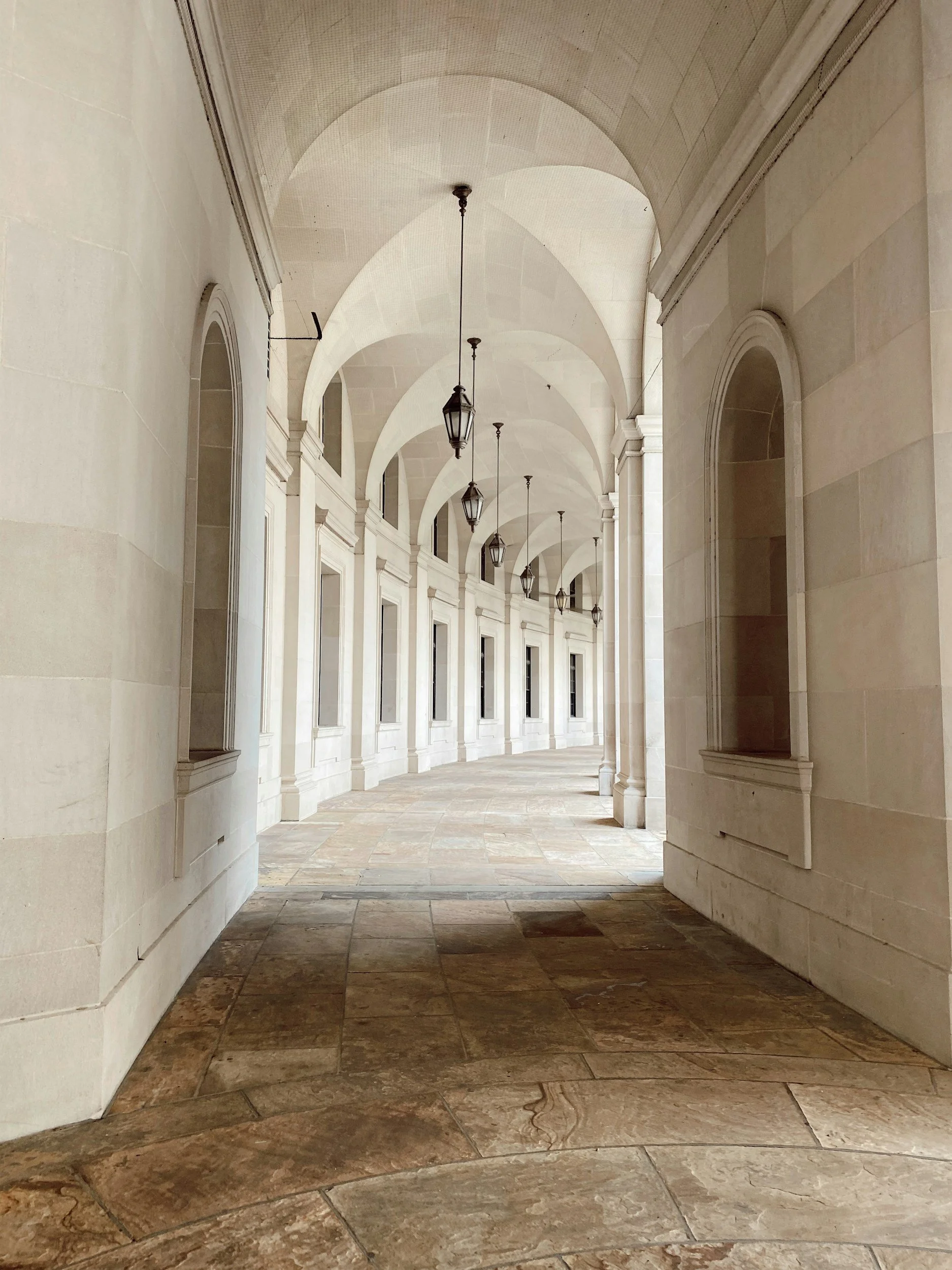 A corridor with arched ceilings, hanging lanterns, and large windows along the side. The walls are made of light-colored stone, and the floor is tiled with large, patterned stone tiles.